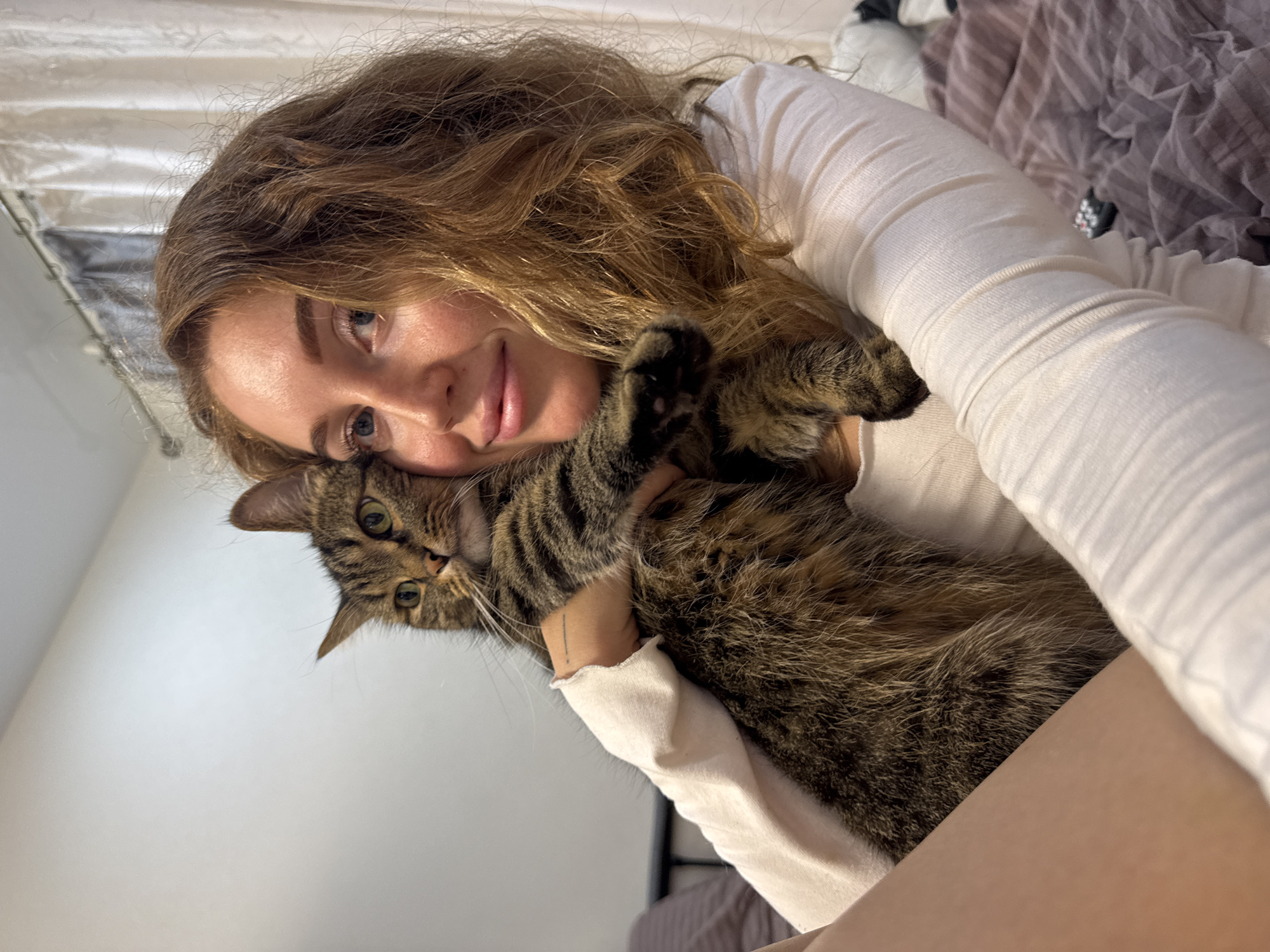 The image presents a cozy indoor scene featuring a young woman and a tabby cat, captured with a slight clockwise rotation. The woman, with long, wavy light brown hair, wears a white long-sleeved top. Her face shows a content and affectionate expression, with her eyes open in the main view, conveying a moment of tenderness.
A tabby cat, distinguished by its brown and black striped fur and green eyes, is being cuddled closely by the woman, its body nestled against her. One of the cat's paws is visible, resting near the woman's hair in a close-up crop, highlighting the intimate interaction and suggesting a strong bond between them. The cat appears calm and comfortable in her embrace.
The background elements indicate an indoor setting, likely a bedroom. Rumpled bedding, including a light purple-brown textured blanket and possibly a white sheet, is visible behind the subjects. A light-colored wall forms part of the backdrop, and a dark, possibly metal, bed frame or headboard is partially seen. The soft, bright lighting suggests either daytime or a well-lit interior. No visible text is present in the image. The overall mood is one of warmth, comfort, and affection, located in Chaikovsky, Russia.