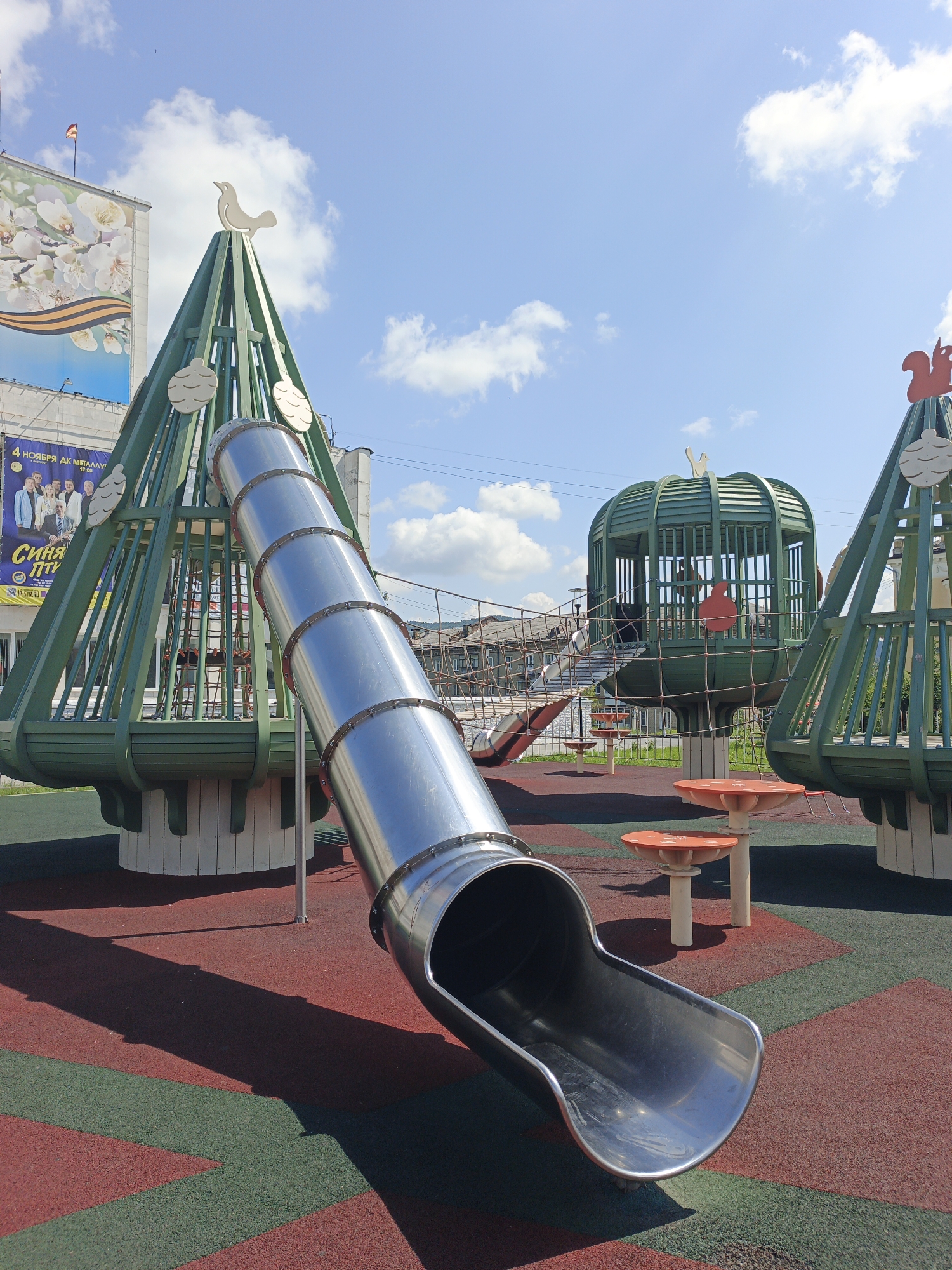 This is an outdoor image of a playground in the city of Zlatoust, Russia, on a sunny day with a blue sky and scattered clouds. The foreground features a shiny, metallic tube slide curving downwards from a green, tree-shaped play structure. The playground surface is a rubberized material with alternating red and green rectangular sections.
In the middle ground, there are several green, cage-like play structures that resemble trees. One structure in the center is cylindrical and has a rounded roof. Red, mushroom-shaped tables are visible near the base of these structures.
In the background, a large building with a billboard advertisement is visible. The billboard features a floral pattern and text in Russian, including "4 НОЯБРЯ ДК МЕТАЛЛУРГ" (November 4th Palace of Culture Metallurg) and a date and time. Another building and some trees are also discernible in the distance. There are no people in the frame. The overall impression is a well-maintained, modern playground designed with naturalistic elements.