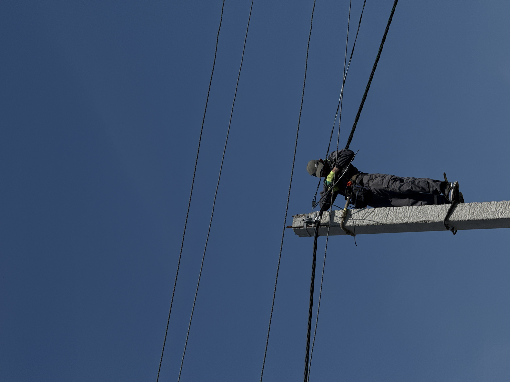An eye-level, wide shot shows a person working on a high-rise electrical tower. The person, dressed in dark clothing and wearing a grey cap and yellow gloves, is lying prone on a horizontal beam, seemingly working on the electrical wires that surround them. The beam itself is weathered and appears to be made of concrete or a similar material, with metal fasteners visible. The background is a clear, deep blue sky, devoid of clouds or other visual distractions. The electrical wires stretch vertically against the sky, framing the worker and the beam. The location in the city of Unknown, Unknown, is not evident from this image.