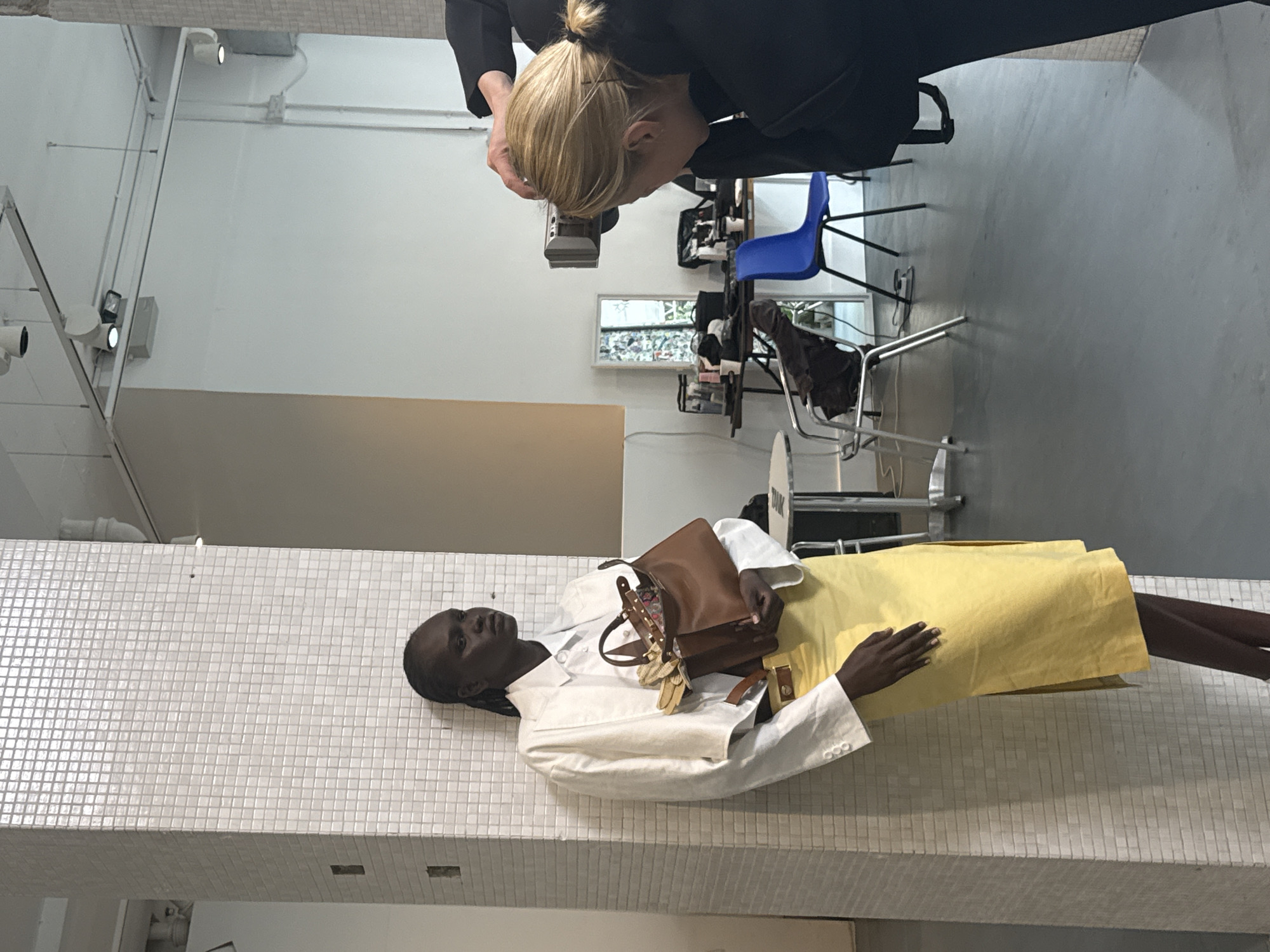 The image shows an indoor scene with a Black woman lying on a tiled platform, likely a display or bench, wearing a white jacket and a yellow skirt. She is holding a brown handbag with decorative elements. Another person, a blonde woman, is taking a picture of her with a camera. The background includes a white wall with pipes and lighting fixtures, a beige wall, and a gray floor. There are chairs and other equipment visible in the background, suggesting the location is possibly a photography studio or fashion-related workspace. Though not explicitly visible, based on the context of fashion photography and studio environments, this image may have been taken in London, United Kingdom.