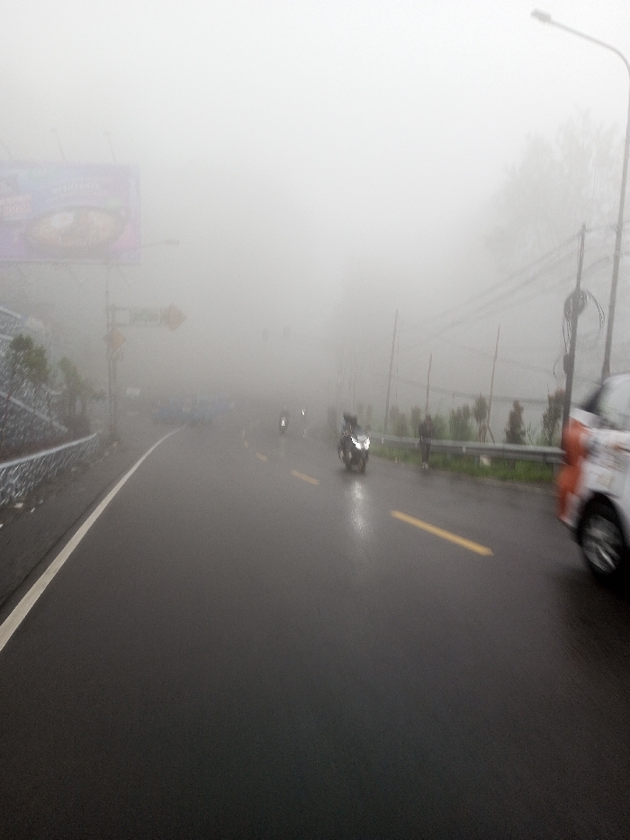 A foggy road scene shows limited visibility. A billboard is visible on the left side of the road. Several motorbikes are seen riding on the road, with a car visible on the right. The road is wet, suggesting recent or current rain. Yellow lane markings are present. Utility poles and a roadside barrier are visible on the right. The heavy fog obscures distant features.