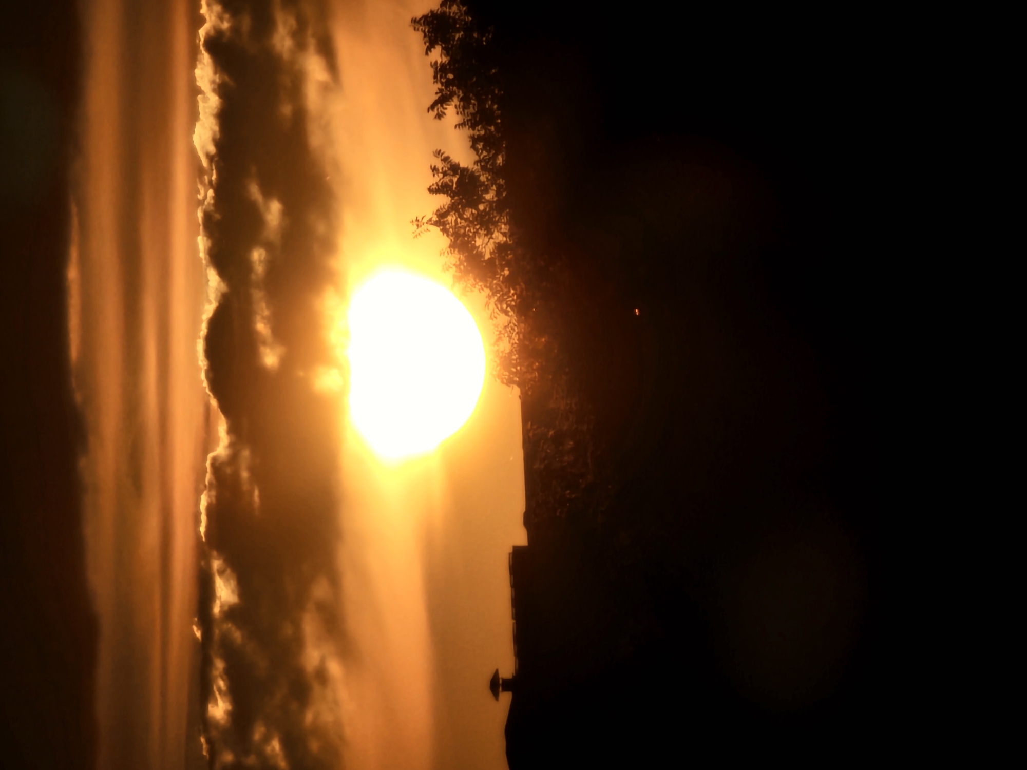 The image shows a sunset over Khimki, Russia. The sun is a bright, white disk surrounded by orange and yellow clouds. In the foreground, there is a silhouette of trees and a building with a cupola. The overall scene is one of a peaceful evening.