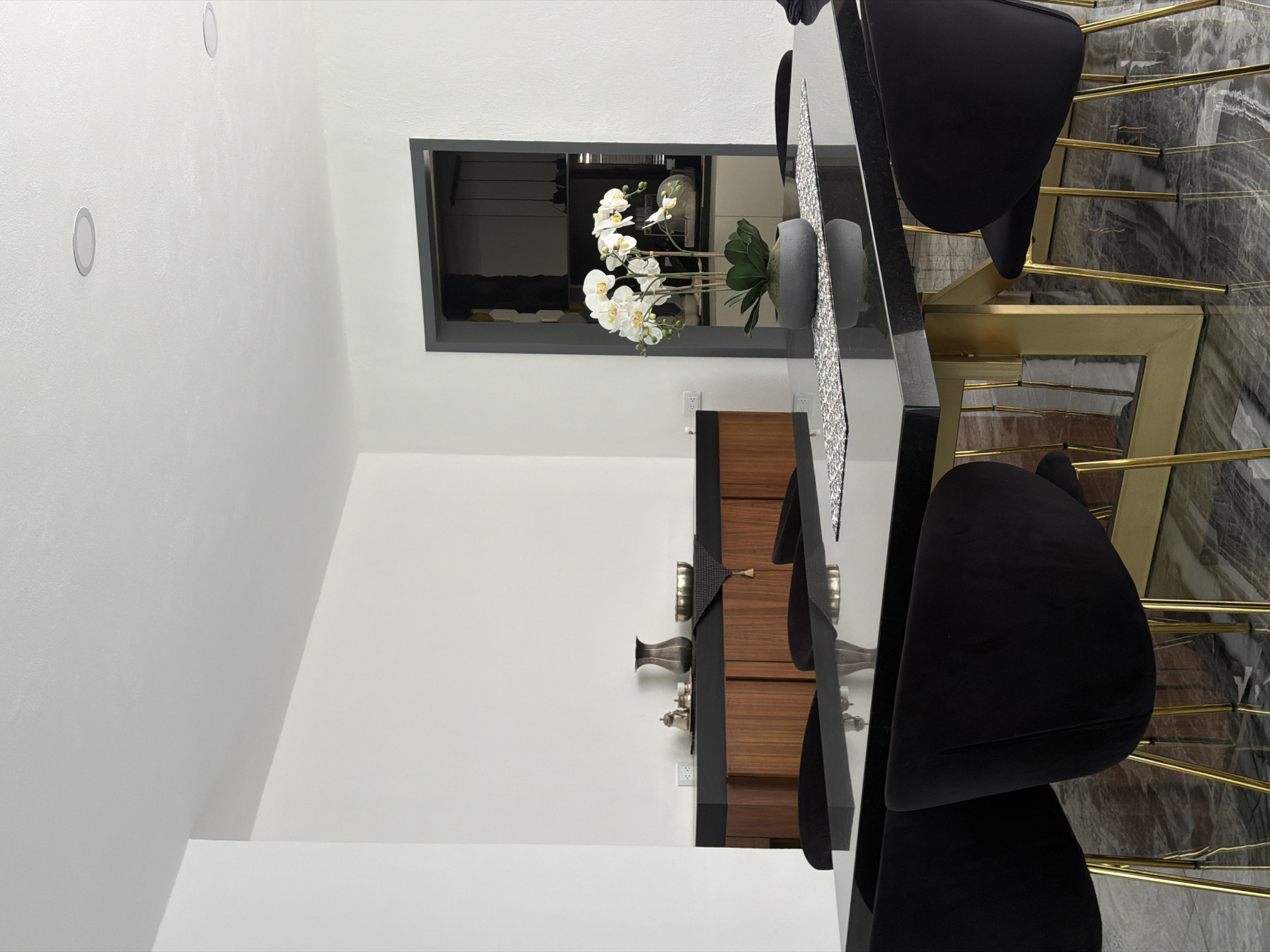 The image shows a contemporary interior, likely a dining area. The scene is dominated by a long, black dining table with gold-colored legs. Black velvet chairs are arranged around the table. A long, silver-colored runner decorates the table surface, along with decorative objects, including a gray vase and white orchids. In the background, a doorway frames a view into another room, reflecting the interior space. A wooden cabinet with dark trim is visible along the wall. The walls are white, with recessed lighting fixtures on the ceiling. The floor appears to be marble or a marble-like material. No people are visible. The location within the city of Puebla, Mexico is not identifiable from the image.
