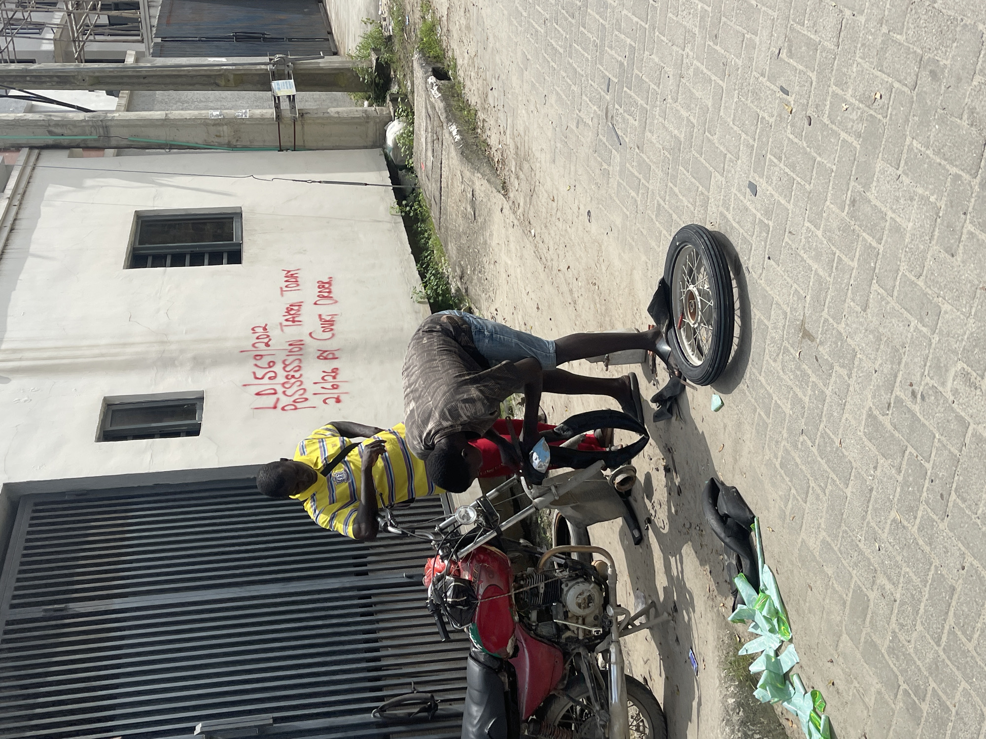 In Igboefon, Nigeria, two men are engaged in an activity with a red motorcycle. One man, wearing a yellow and blue striped shirt, stands beside the motorcycle, looking down. The other man, dressed in a dark patterned shirt and denim shorts, is actively working on the motorcycle's front wheel, which appears to be detached and lying on the ground. The front tire and inner tube are also on the ground, with some green plastic material nearby. The scene is set on a paved street with a building in the background that has red graffiti on its wall. The graffiti reads "LD/569/2012 POSSESSION TAKEN TODAY 2/6/26 BY COURT ORDER."
