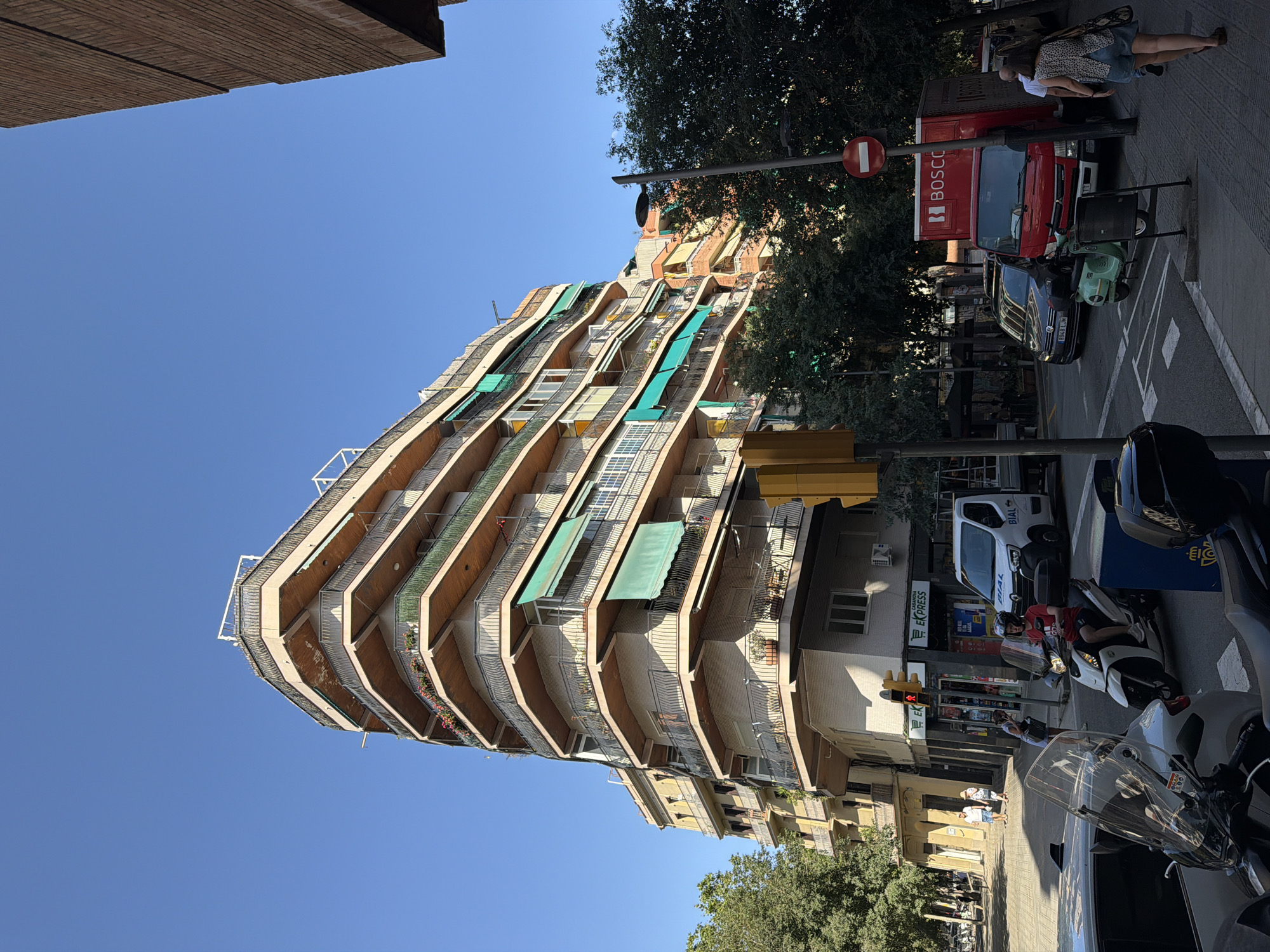 The image captures a street scene in Barcelona, Spain, under a clear blue sky, suggesting a sunny day. The dominant subject is a distinctive, curved apartment building with numerous balconies featuring metal railings and green awnings. These balconies create a wave-like pattern along the building's facade.
On the street below, several objects and people are visible. A red truck with "BOSCO" written on its side is parked. A dark Volkswagen car and a light green scooter are also present. A few individuals are seen on the sidewalk, including a couple standing near the red truck, and a person walking. Further down the street, another scooter and a white van with "BIAL" written on it are parked in front of a store. Traffic lights and street signs are also visible.
The overall impression is of a typical urban environment in Barcelona, with a focus on the unique architecture of the apartment building. The sunlight casts shadows, indicating it's likely daytime. There are no overt displays of emotion, and the scene appears to be a snapshot of daily life on a sunny day.