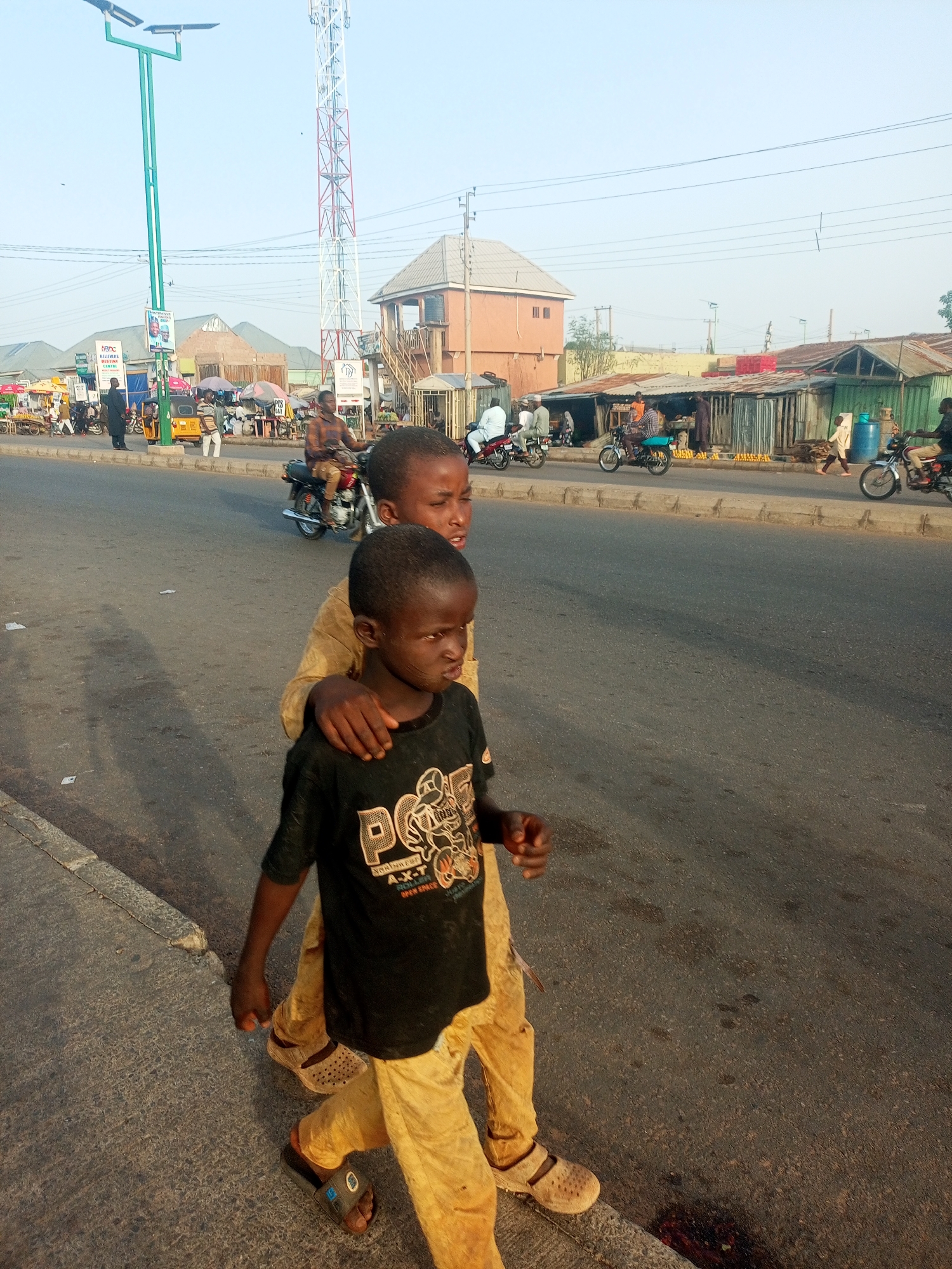 Two young boys are walking along the edge of a road in Tumfure, Nigeria. The older boy has his arm around the younger boy's shoulders. Motorbikes are driving on the road. Shops are visible in the background. A communication tower is visible in the background.