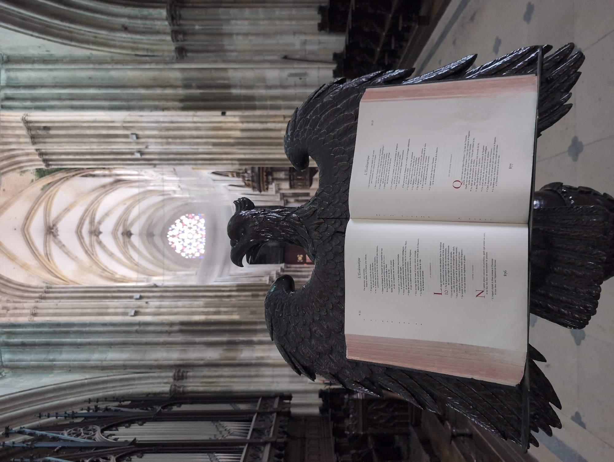 The image shows a view inside a cathedral, likely in Le Tréport, France, focusing on an ornate eagle-shaped lectern holding an open book. The lectern is dark wood and intricately carved, with spread wings forming the base. The book is open to two pages of text, printed in French. The background reveals the grand architecture of the cathedral, with tall columns, arched ceilings, and a rose window visible in the distance. Sunlight filters through the stained glass, casting colorful patterns. The overall atmosphere is one of solemnity and historical grandeur.