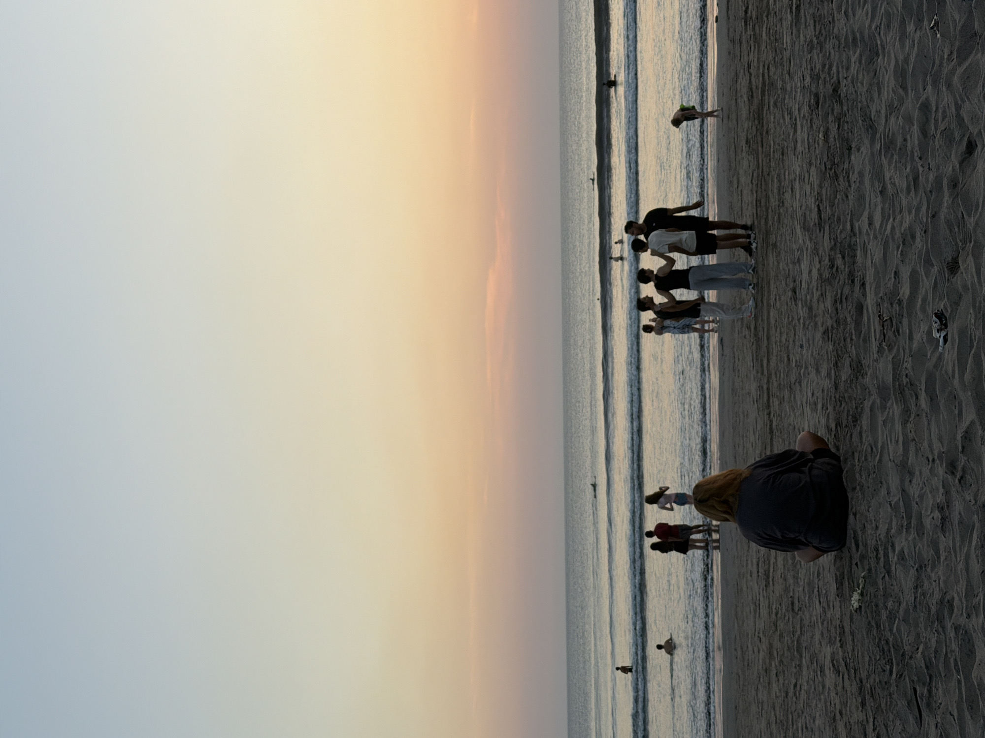 The image depicts a sunset scene at Kuta Beach in Bali, Indonesia. The foreground is dominated by dark, rippled sand, with a solitary person sitting with their back to the camera, facing the ocean. This individual has blonde hair and is wearing a dark grey t-shirt and shorts.
In the middle ground, a group of five young people stands near the water's edge, appearing to be engaged in conversation. Their postures suggest a casual gathering. Further out in the ocean, several other individuals are visible. Some are wading in the shallow water, while others are further out, with one person on a surfboard. The ocean is calm with gentle waves rolling towards the shore.
The background is a vast expanse of the ocean meeting the sky. The sky displays a gradient of soft colors characteristic of a sunset, transitioning from a pale blue at the top to hues of peach and pink closer to the horizon. The overall atmosphere is peaceful and serene, capturing a typical evening at a popular beach destination.