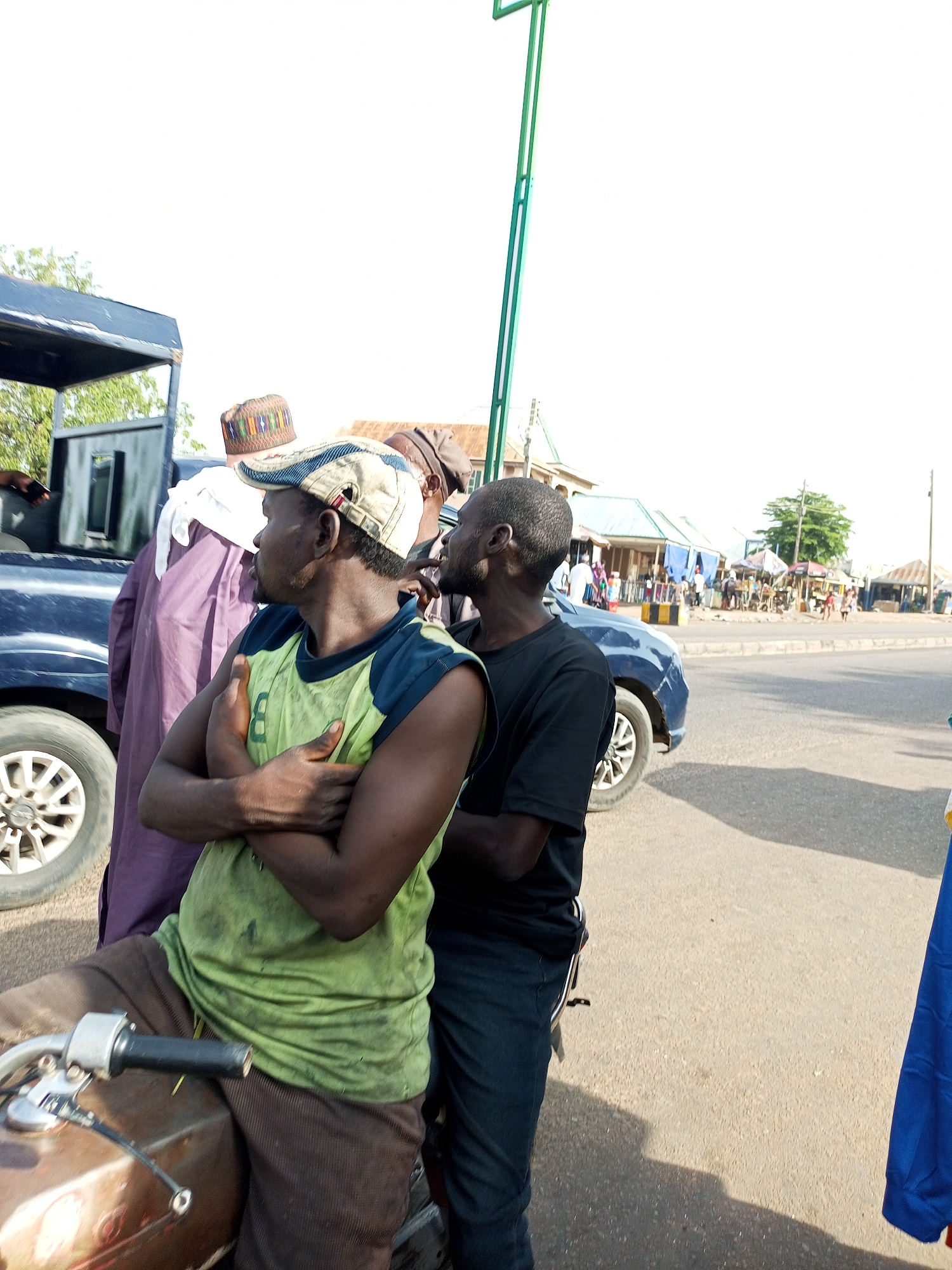The image depicts a street scene in Gombe, Nigeria, with several individuals and vehicles. In the foreground, two men are the primary subjects. The man on the left, wearing a light green sleeveless t-shirt and brown pants, is seated on what appears to be a motorcycle. He has his arms crossed and is looking to the right. He wears a distinctive cap. To his right stands another man in a black t-shirt and dark pants, also looking towards the right.
Behind these two men, a dark-colored vehicle, possibly a truck or SUV, is partially visible. A person in a purple robe and a white head covering stands next to it. Further in the background, a busy street scene unfolds with several small shops or stalls lining the road. People are milling about, and various vehicles, including cars and what might be market stalls on wheels, are present. The sky is bright and overcast. A tall, green pole is a prominent vertical element in the background, possibly part of a street light or a sign. The overall environment suggests a bustling urban or semi-urban setting with commercial activity.