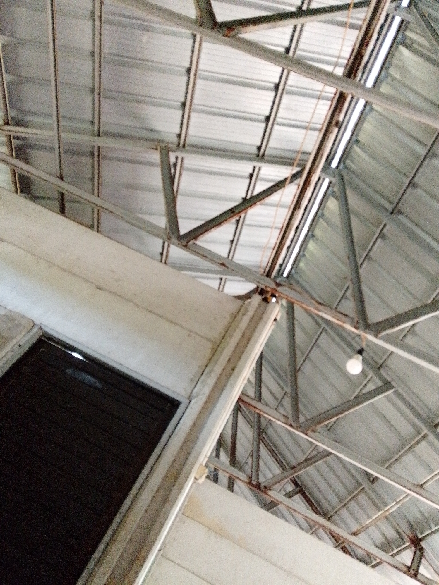 The image, taken from a low angle, shows the interior of a building in Bukit Ketok, Indonesia. The ceiling is composed of corrugated metal sheets supported by a network of metal beams. A single light bulb hangs from the beams. A portion of a wall, painted white, is visible in the foreground, with a dark, rectangular opening within it, possibly a doorway or window.