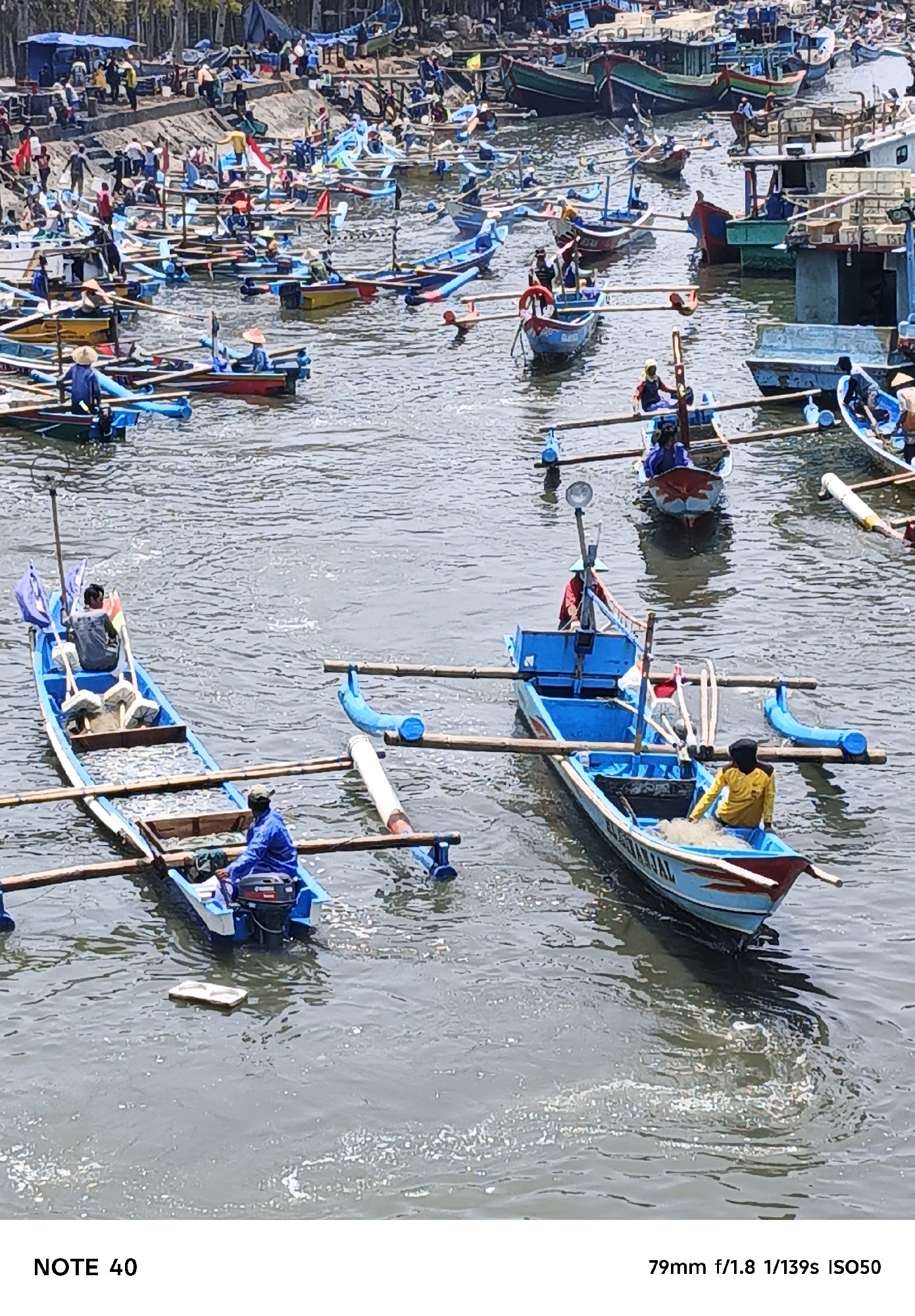 A high-angle medium shot captures a busy harbor scene in Kelurahan Tegalkamulyan, Cilacap, Indonesia, with numerous traditional fishing boats of varying sizes and colors dotting the choppy water. The boats are primarily wooden, painted in vibrant blues, reds, yellows, and greens, and many are equipped with outriggers and oars. Several individuals are visible, predominantly men dressed in casual clothing, some wearing hats, actively engaged in boat-related activities. One person in a blue shirt and hat operates a small outboard motor on a boat in the foreground. Another individual in a blue jacket is seated in a blue and red boat in the middle ground, while a person in a yellow shirt and black head covering is seen tending to nets in a blue and red boat. People and structures are visible on the shore in the background, suggesting a bustling marketplace or landing area. The water appears murky and somewhat polluted, with scattered debris visible on the surface. The overall impression is one of a lively and active fishing community.