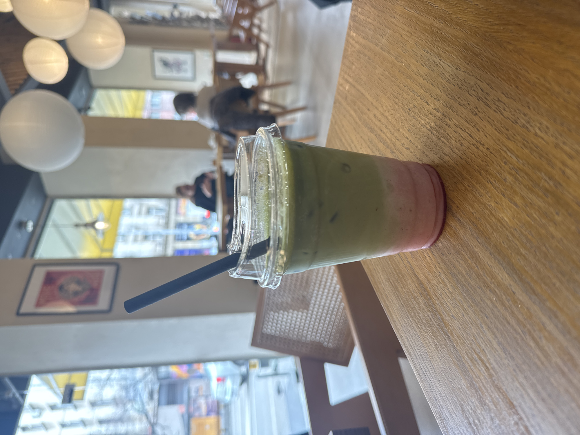 The image shows a plastic cup with a layered drink, likely matcha and strawberry, with a black straw on a wooden table inside a cafe. The cafe appears to be located in Sofia, Bulgaria, based on the distinctive architecture and streetscape visible through the window. Customers are seated in the background, and spherical light fixtures hang from the ceiling.
