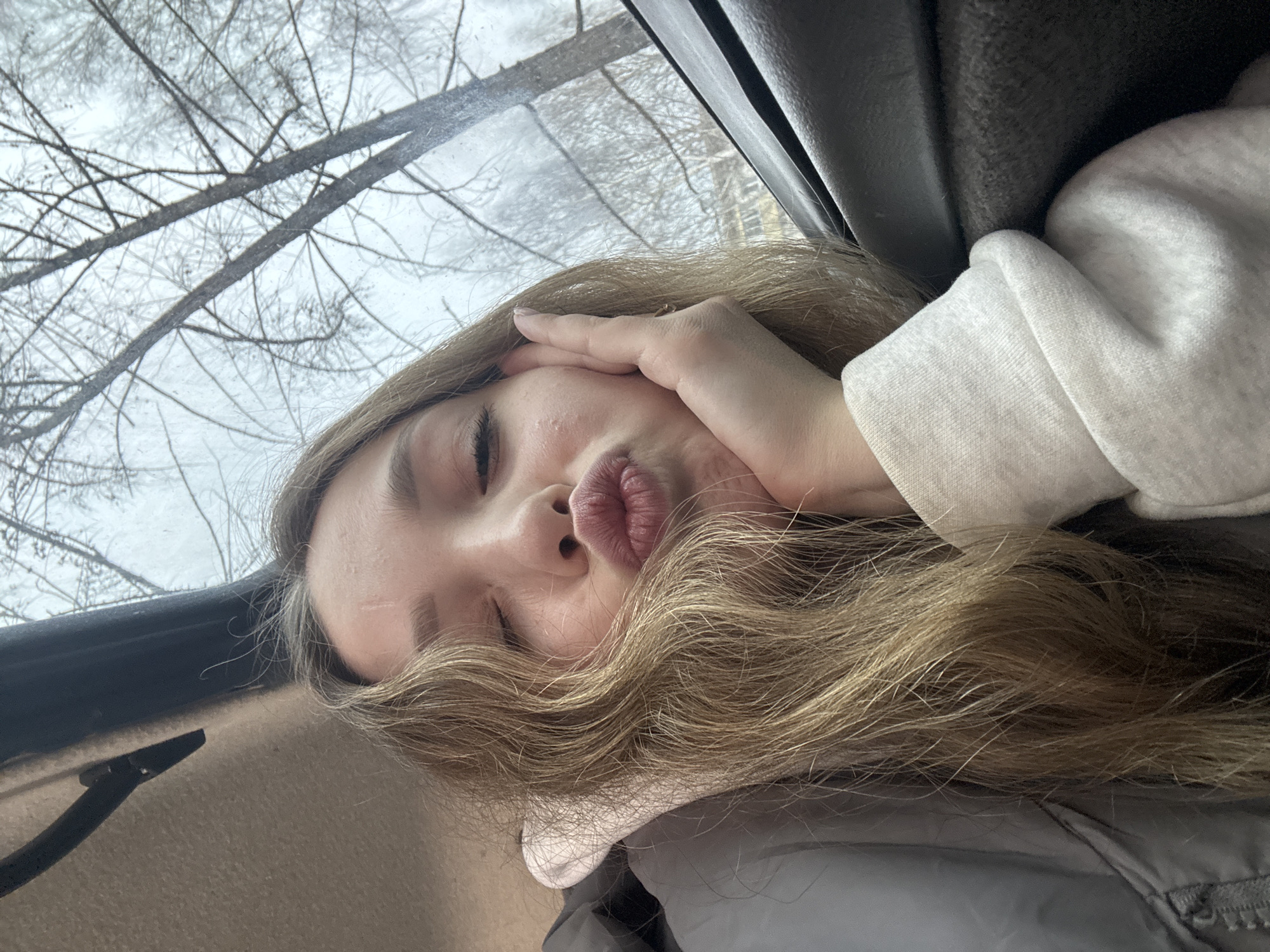 A young woman is photographed in what appears to be a car interior. She has fair skin and long, blonde hair that covers part of her face. She is making a pouting expression with her lips and her right hand is touching her face, covering her forehead. She is wearing a white hoodie or sweater. Behind her, visible through the car window, are bare tree branches against a white sky, suggesting a winter scene. The interior of the car features a beige or light brown color scheme with dark gray accents. There is no specific information in the image that definitively identifies the location as Izhevsk, Russia.