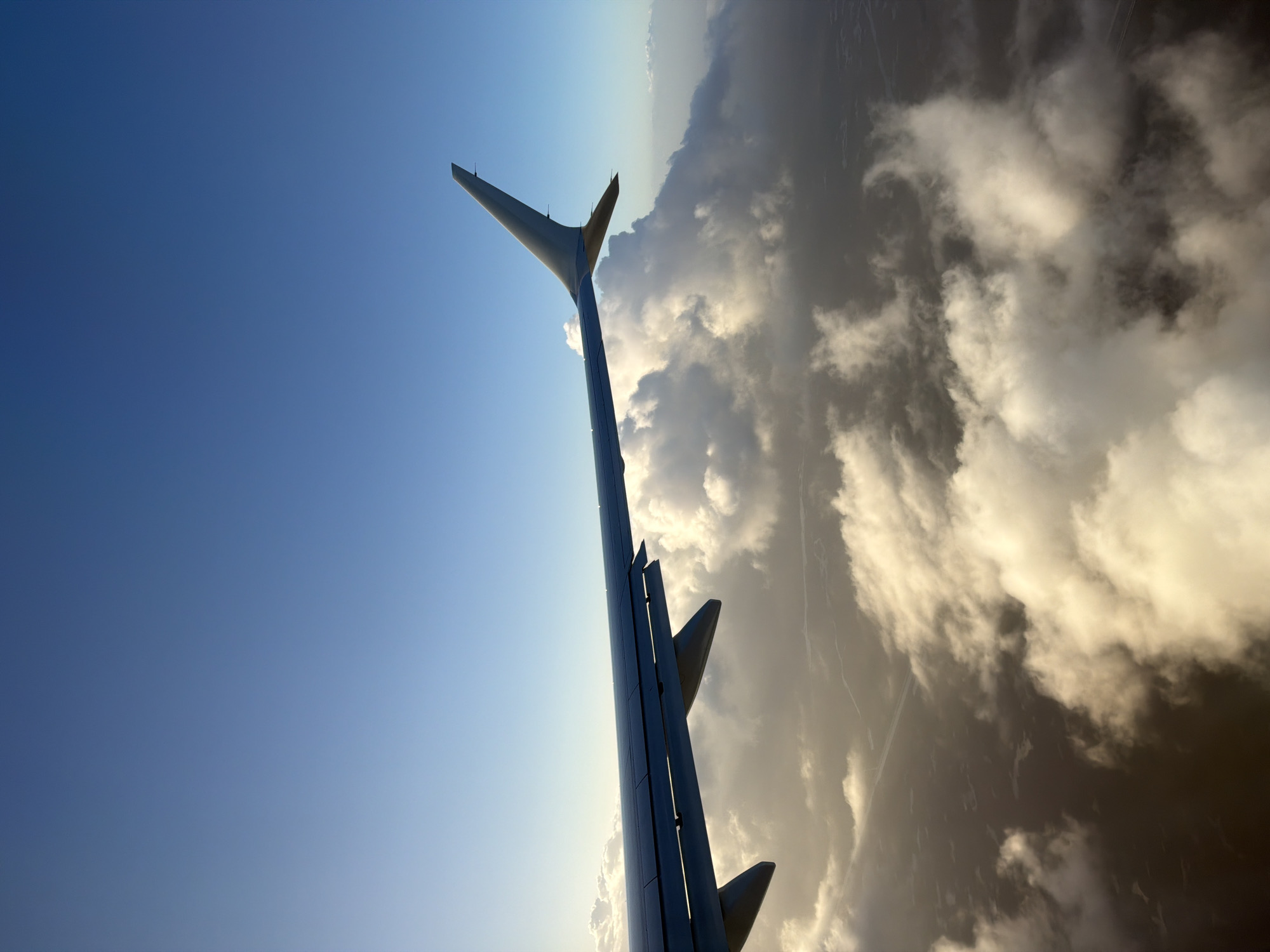 The image shows an airplane wing and tail fin extending against a backdrop of blue sky and clouds. The wing and tail fin are silver or gray in color. The sky transitions from a clear blue to a lighter hue near the horizon. The clouds vary in density, some appearing as puffy formations and others as darker, shadowy masses. The location is not evident in the image. The city, state, and country is Unknown, United States of America.
