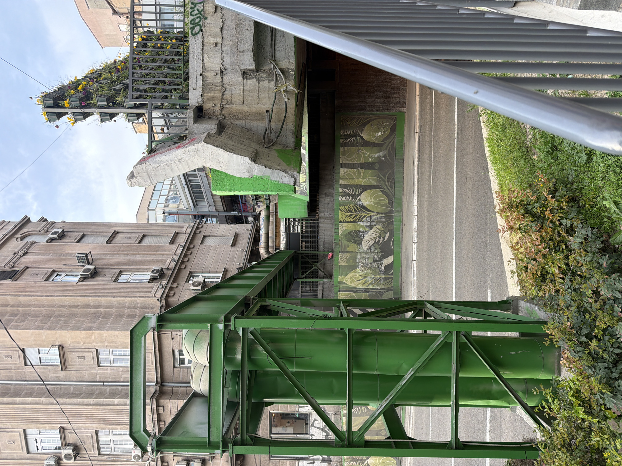 The image depicts an urban scene in Belgrade, Serbia, viewed from an elevated perspective. A series of large, green, industrial pipes are supported by a matching green metal framework. Adjacent to the pipes is a multi-story building with a beige facade, adorned with numerous windows and air conditioning units. The ground beneath the pipes is partially a street with white lines, and partly foliage with green plants. A concrete structure with remnants of green paint and graffiti, possibly part of a bridge or overpass, looms overhead. In the background, a building features decorative flowers on its upper levels. A metal railing, partially obscured, occupies the foreground.