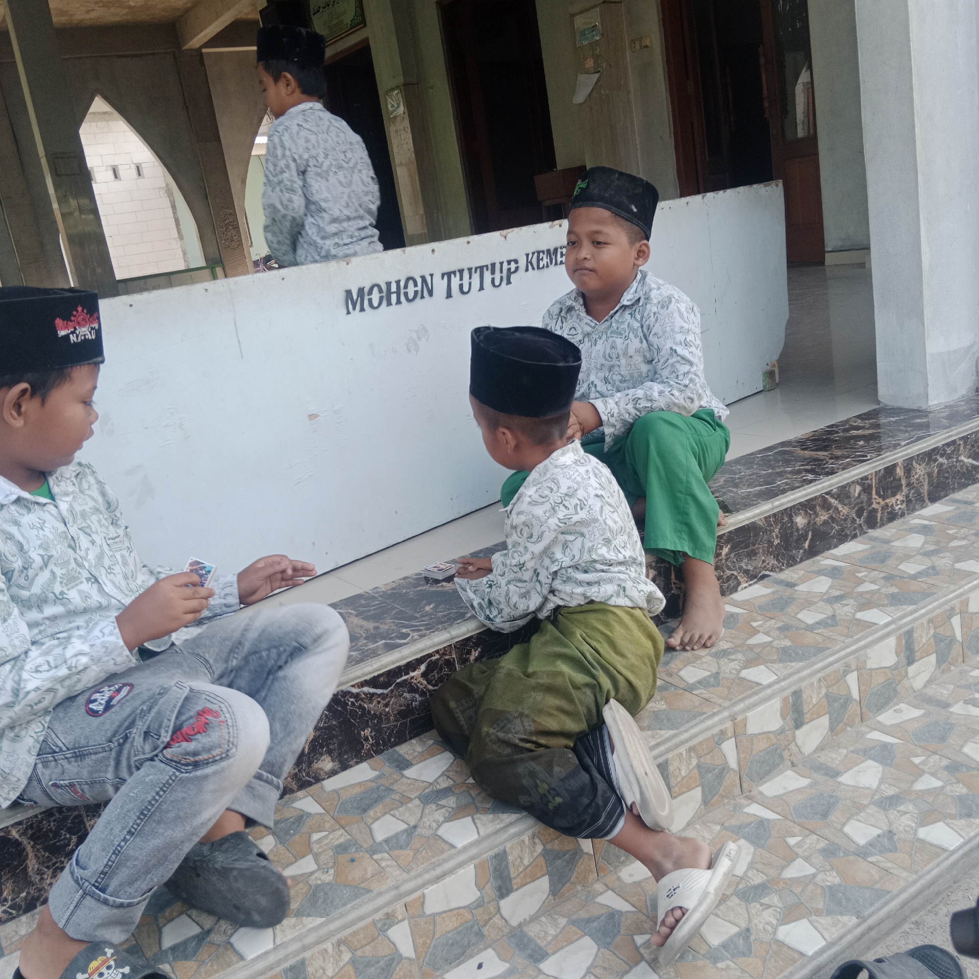 In the city of Tegowanu Kulon, Indonesia, three young boys are seated on the tiled steps of a building. The boy on the left is wearing a black skullcap with red insignia, a white patterned shirt, and grey jeans. He is holding a small card in his right hand and appears to be looking at it. The boy in the center, also wearing a black skullcap and a white patterned shirt, is seated with his legs crossed and is looking downwards. The third boy, to the right, has a black skullcap and is wearing a white patterned shirt and green pants. He is looking to his left. Behind the boys is a white wall with dark lettering that reads "MOHON TUTUP KEME." The environment suggests an outdoor area, possibly near a place of worship or a public building, with tiled flooring and architectural elements visible.