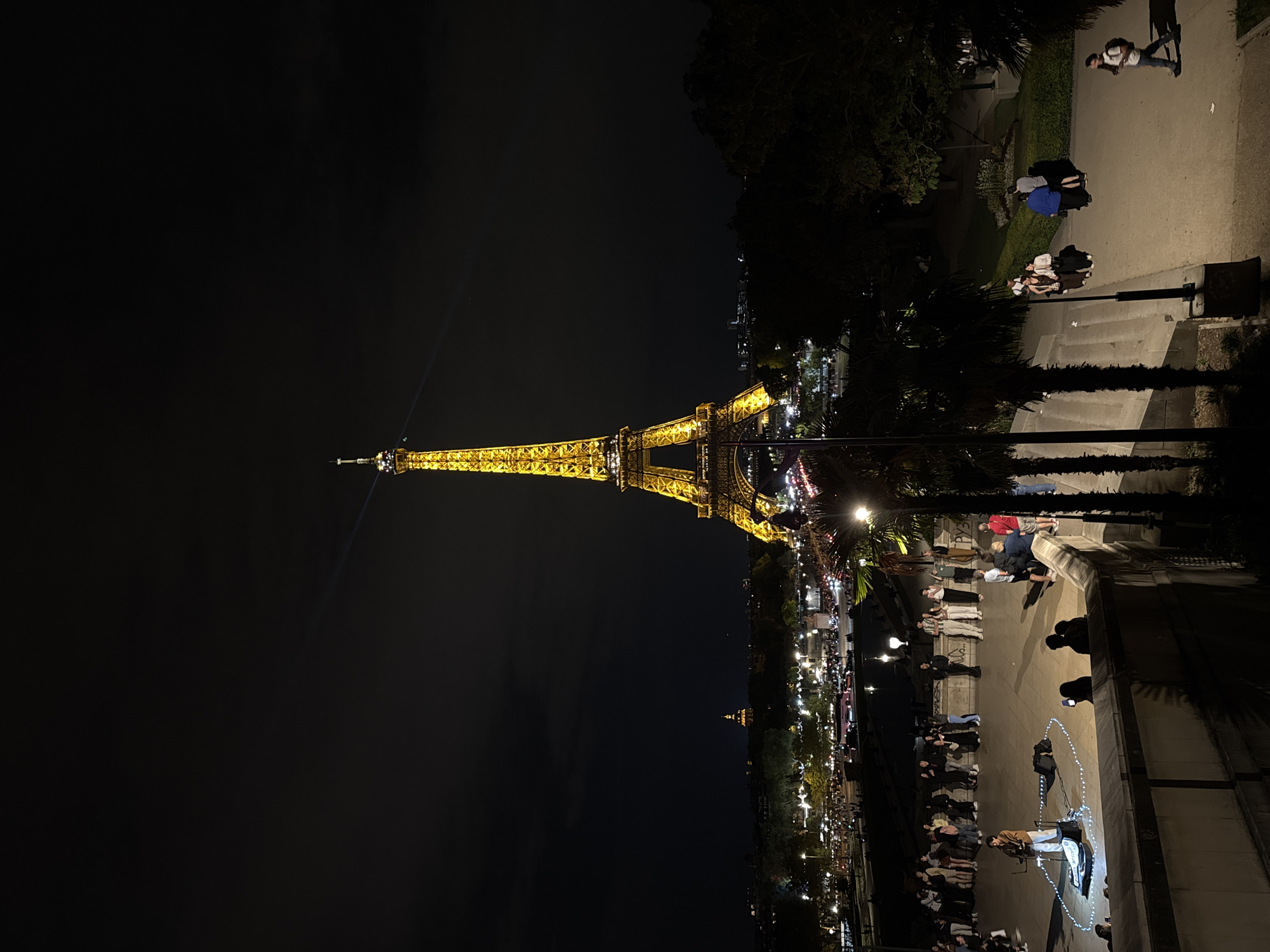 The image shows the Eiffel Tower in Paris, France, at night. The tower is illuminated and a beam of light is visible from the top. The ground level is partially visible, showing a crowd of people near the tower. Streetlights and building lights are visible in the background. A walkway with people is seen on the right side of the image. The sky is dark.