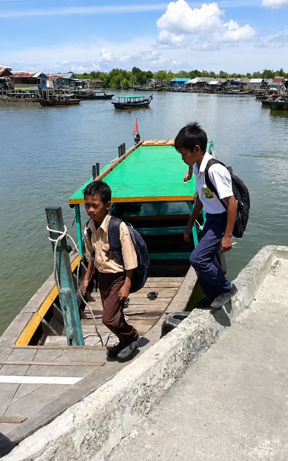 Two young boys, wearing school uniforms and backpacks, are disembarking from a wooden boat docked at a concrete pier. The boat has a green-painted upper deck and wooden planks forming its flooring. The boy in the foreground, on the left, wears a light brown shirt and brown pants, carrying a dark backpack. The boy on the right wears a white shirt and blue pants, also with a dark backpack. They are in a waterway, likely a river or canal, with a village of stilt houses visible on the far bank, characterized by colorful roofs and wooden structures. The water is calm and reflects the blue sky with scattered white clouds. The environment suggests a coastal or riverside community in Indonesia, consistent with the provided location of the City of Medan. The overall scene captures a daily commute or activity related to school in this particular setting.
