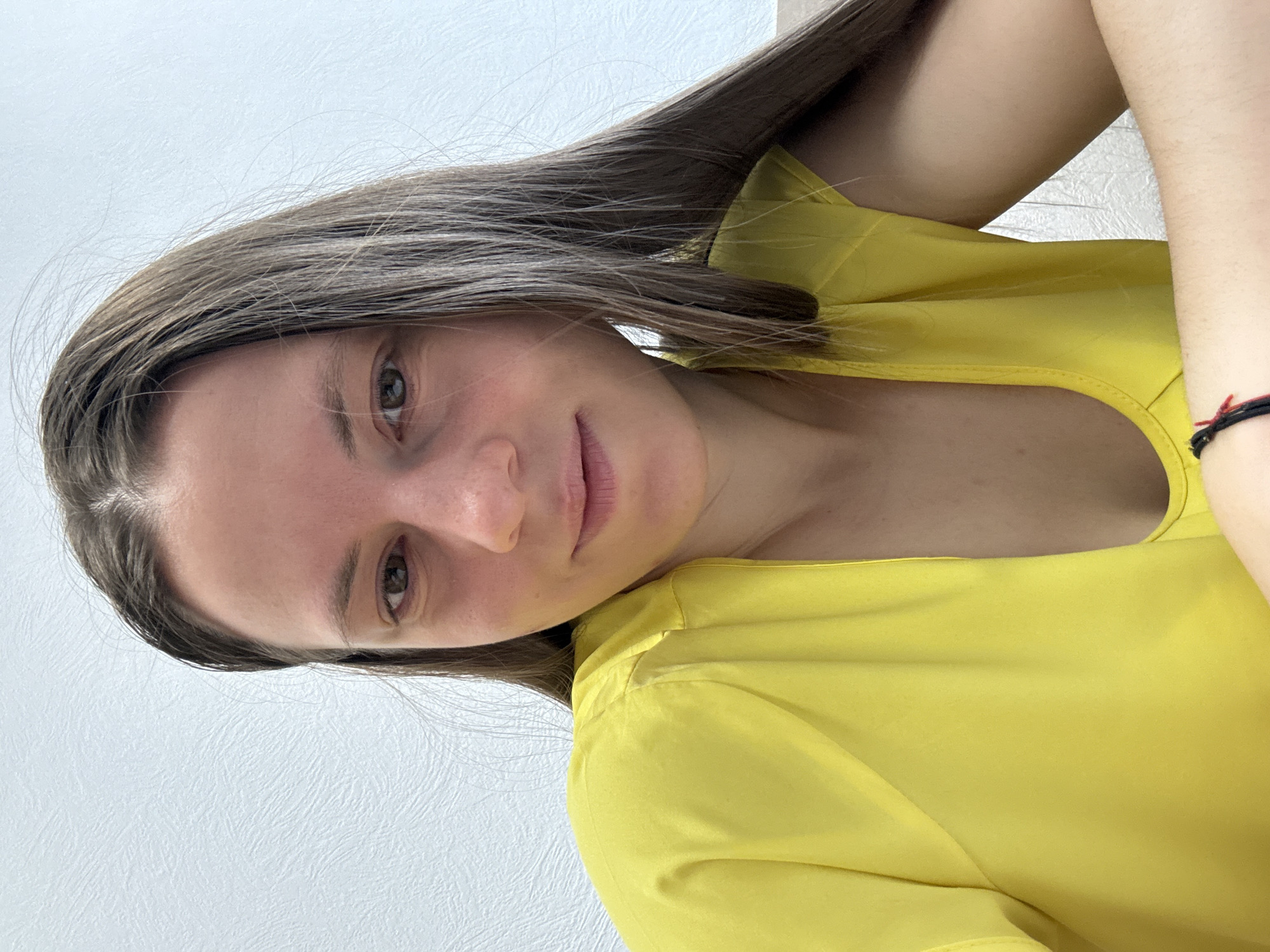 A young woman is indoors with her head tilted to the left. She has brown hair and light skin, and is wearing a yellow top. A white wall is visible in the background. No information about the city of Chaikovsky, Russia is visible.
