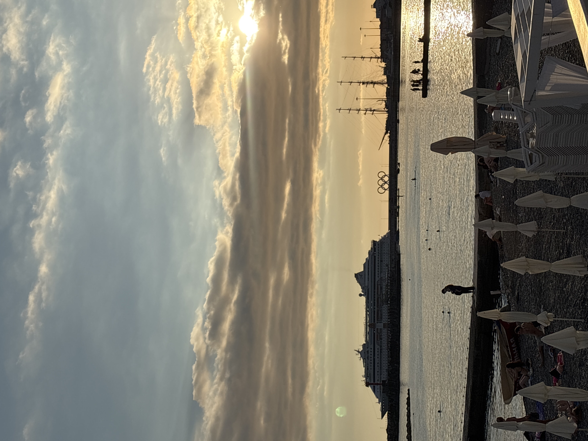 This media file depicts a sunset scene at a beach in Sochi, Russia. The sky is filled with dramatic, golden-hued clouds, illuminated by the setting sun. In the background, several tall-masted sailing ships are docked, silhouetted against the warm light. Further out at sea, a large cruise ship is visible. The Olympic rings monument stands prominently in the water, a clear indicator of the location.
On the beach in the foreground, beach umbrellas and chairs are set up, some closed and others open. Several people are scattered on the pebbled shore, relaxing or walking. The water is calm, reflecting the soft colors of the sky. A person is seen standing near the water's edge, and another couple is walking along the pier. The overall atmosphere is peaceful and serene, with the golden hour light casting a warm glow over the entire scene. The time of day is clearly sunset, and the weather appears to be clear with beautiful cloud formations. There is no visible text in the image.