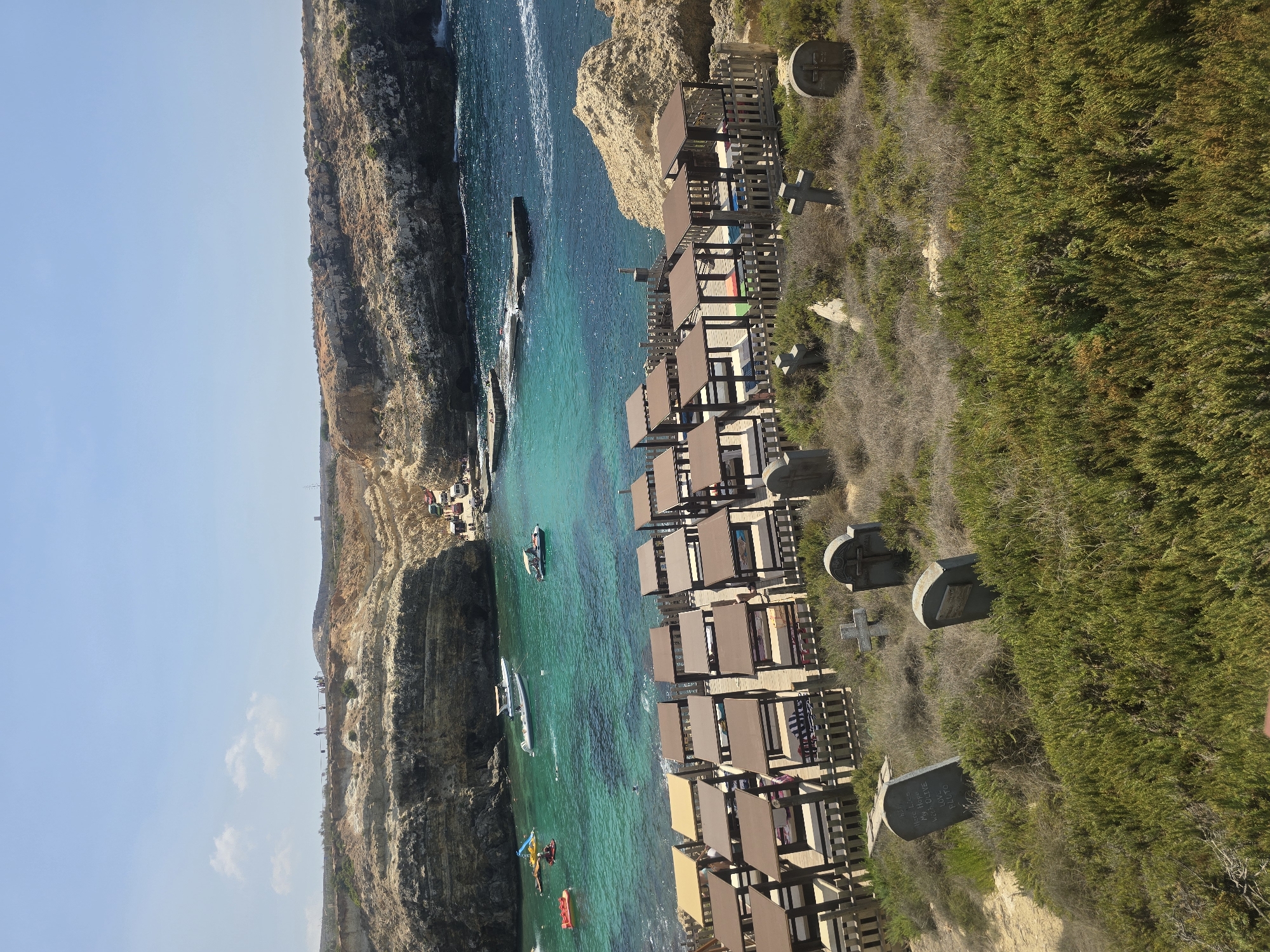 The image depicts a sun-drenched coastal scene in Mellieħa, Malta, likely at a beach resort. In the foreground, a collection of gravestones stands amidst dry, scrubby vegetation, a peculiar and somber element against the bright backdrop. Beyond this, rows of covered lounge chairs, arranged in orderly fashion, suggest a leisure area. The turquoise waters of the sea are visible, dotted with various boats, including a small yacht and a few recreational vessels, indicating people are engaging in water activities. Rocky cliffs form the backdrop of the bay, with a few structures or platforms visible along the water's edge and on the cliffs above. The sky is clear and blue, with a few wispy clouds, indicative of fair weather. The overall impression is a juxtaposition of leisure and a morbid, perhaps artistic, display.