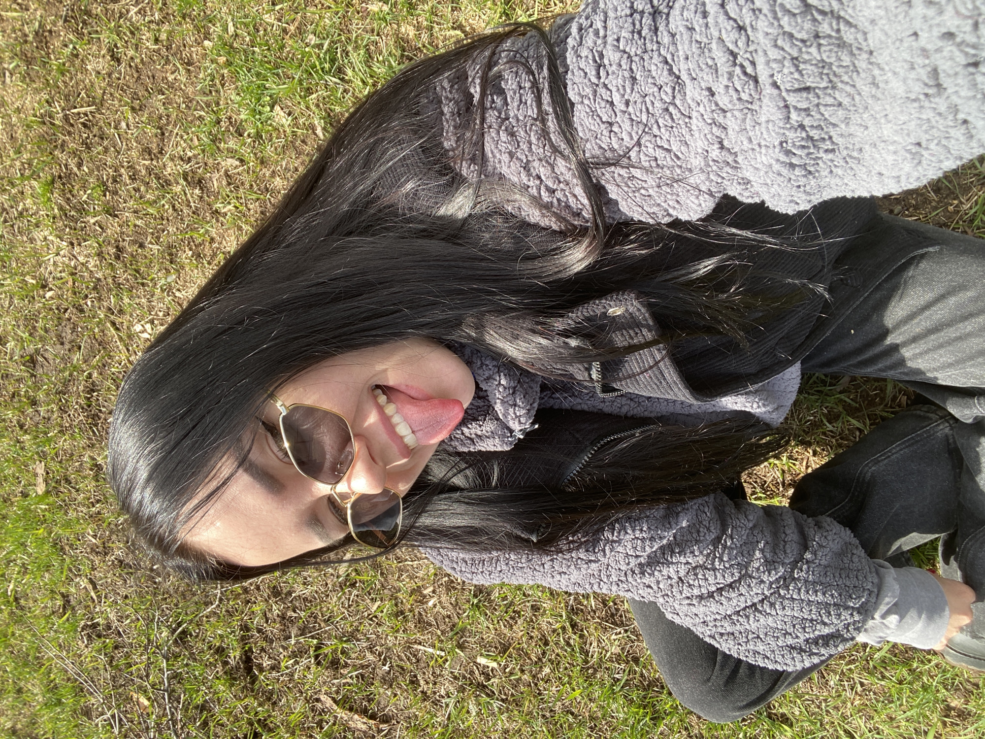 A woman is lying on a patch of grass, seemingly outdoors. She is wearing sunglasses, a gray textured jacket over a dark shirt or jacket, and dark pants. Her long dark hair is spread around her head. She is sticking out her tongue and appears to be taking a selfie. The location is not specified as being in Chaikovsky, Russia. The environment consists of grass, some of which appears dry and brown, suggesting it may be early spring or late fall.