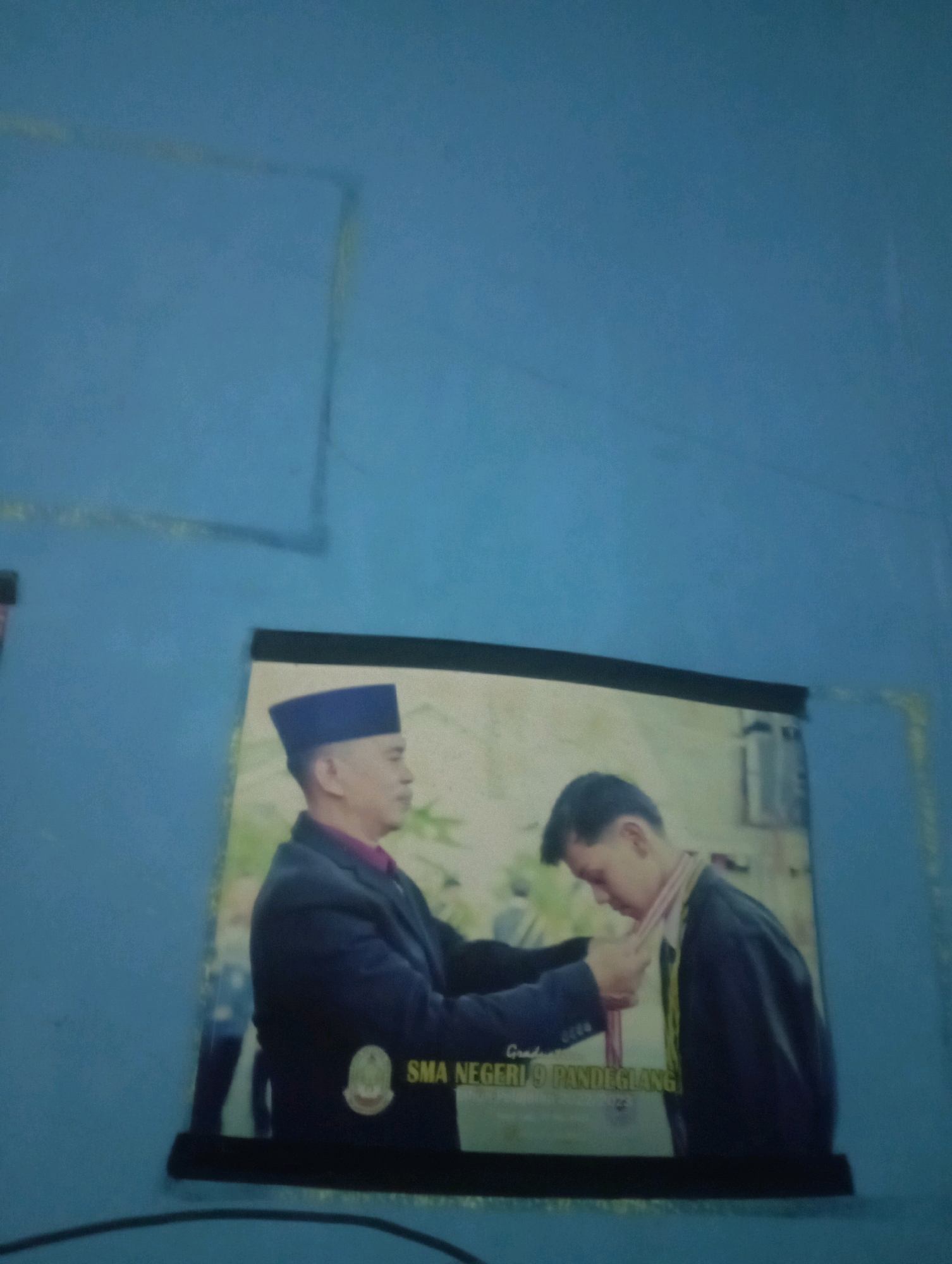 This is a photograph taken indoors, likely in a residential setting in Senangsari, Indonesia. The background consists of a textured, light blue wall with some faint marks or shadows. Affixed to the wall is a framed photograph that occupies the lower half of the image.
The photograph within the frame depicts an awarding ceremony. On the left is a man wearing a dark suit, a purple shirt, and a dark blue cap, appearing to be an official or dignitary. He is in the process of placing a medal or sash around the neck of a younger man on the right. The younger man is wearing a dark academic gown or robe over a light-colored shirt and a decorative sash or stole. His head is bowed slightly, and his expression suggests humility or gratitude.
At the bottom of the framed photograph, text is visible. Partially obscured, it reads "Graduation" and then "SMA NEGERI 9 PANDEGLANG," indicating that the event is a graduation ceremony for SMA Negeri 9 Pandeglang, a high school in Pandeglang, Indonesia. The overall mood of the photograph within the frame is one of accomplishment and recognition. The lighting in the scene is dim, with the primary light source appearing to come from the front of the framed photograph.