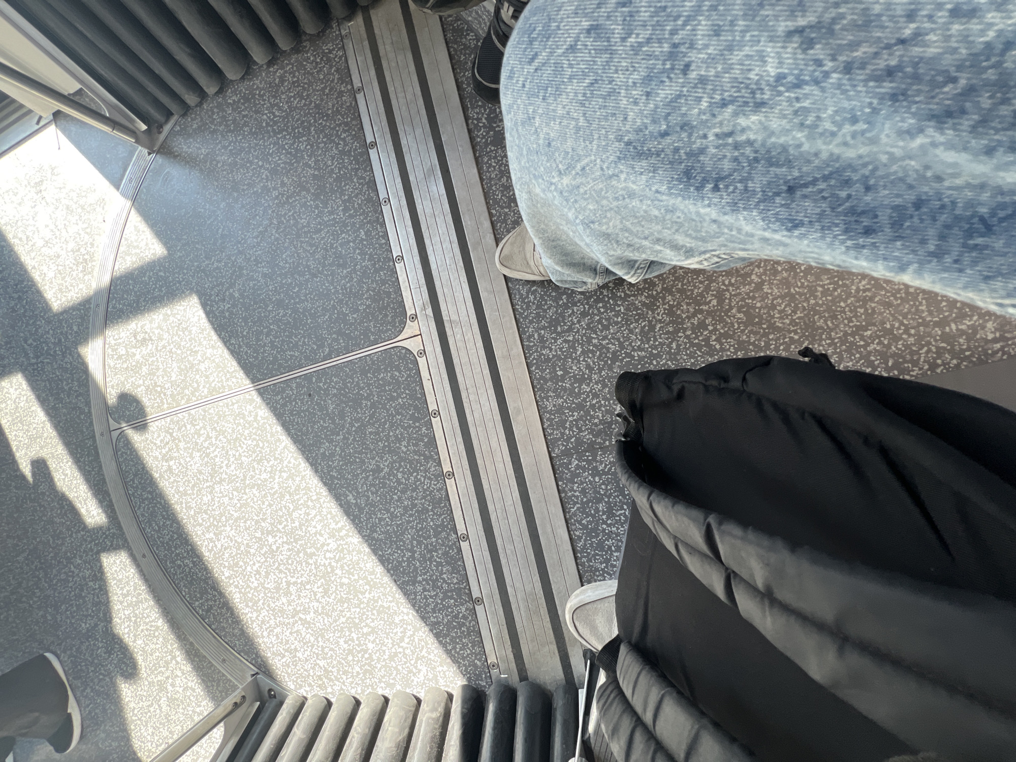The image shows a portion of the interior of a Strasbourg tram, likely a CTS (Compagnie des Transports Strasbourgeois) tram based on the interior design. The floor has a mottled gray pattern with metal strips running along the floor, potentially delineating sections or providing grip. Seating runs along the edge of the image.
A person wearing jeans and shoes is standing in the tram. A black jacket is also present. The lighting is bright, suggesting daytime operation. The focus is on the immediate surroundings within the tram rather than providing a broader view of Strasbourg.