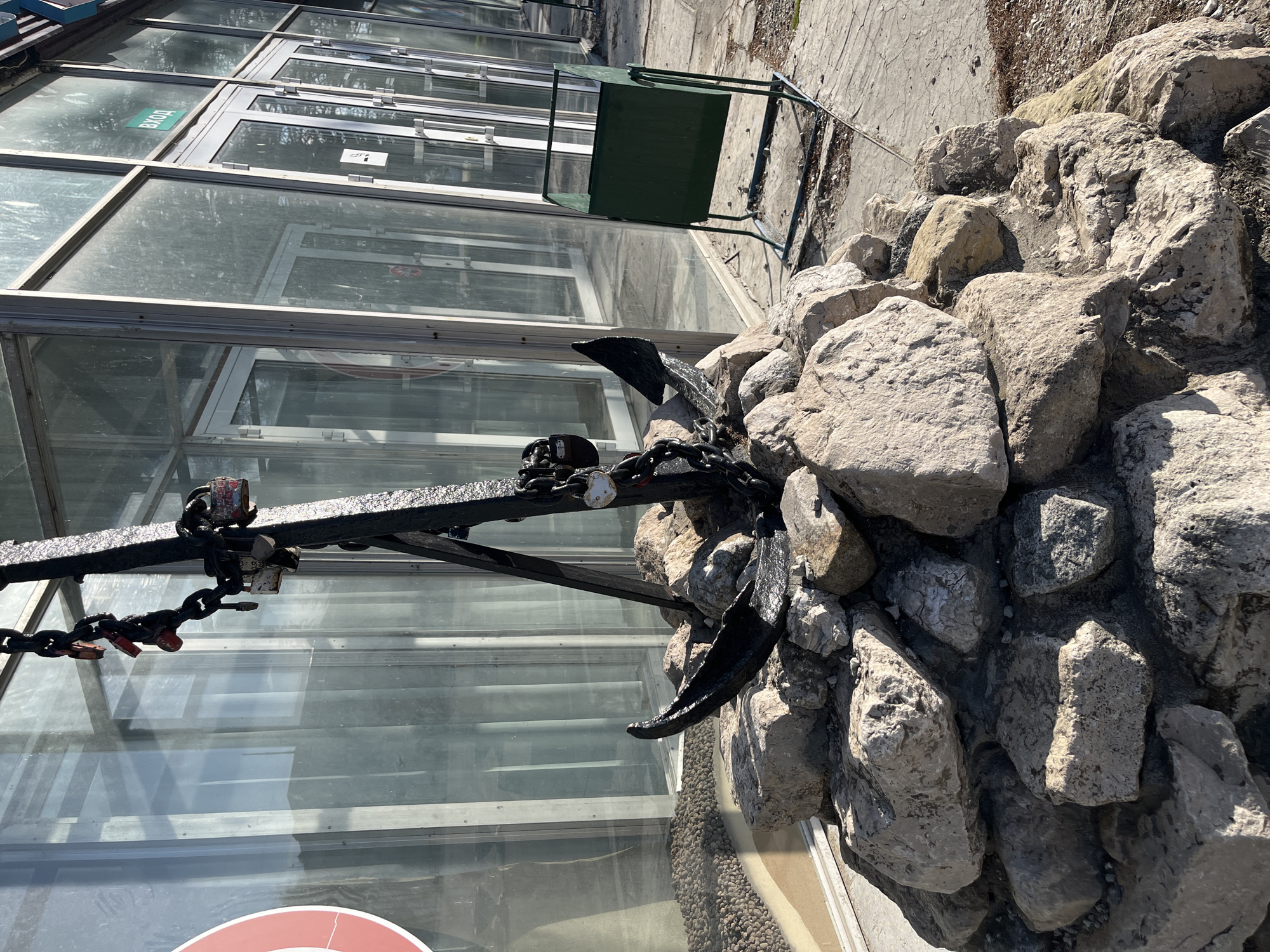 The image depicts a maritime-themed display in Ulyanovsk, Russia, featuring a black anchor prominently situated amidst a pile of gray rocks. The anchor, with visible chains and attached locks, is positioned against the backdrop of a modern glass-walled building, potentially a commercial structure or a greenhouse-like enclosure. The scene suggests a blend of industrial, natural, and maritime elements. A green, metal outdoor chair is partially visible to the left, adding to the urban context. The overall environment appears to be an outdoor public space.