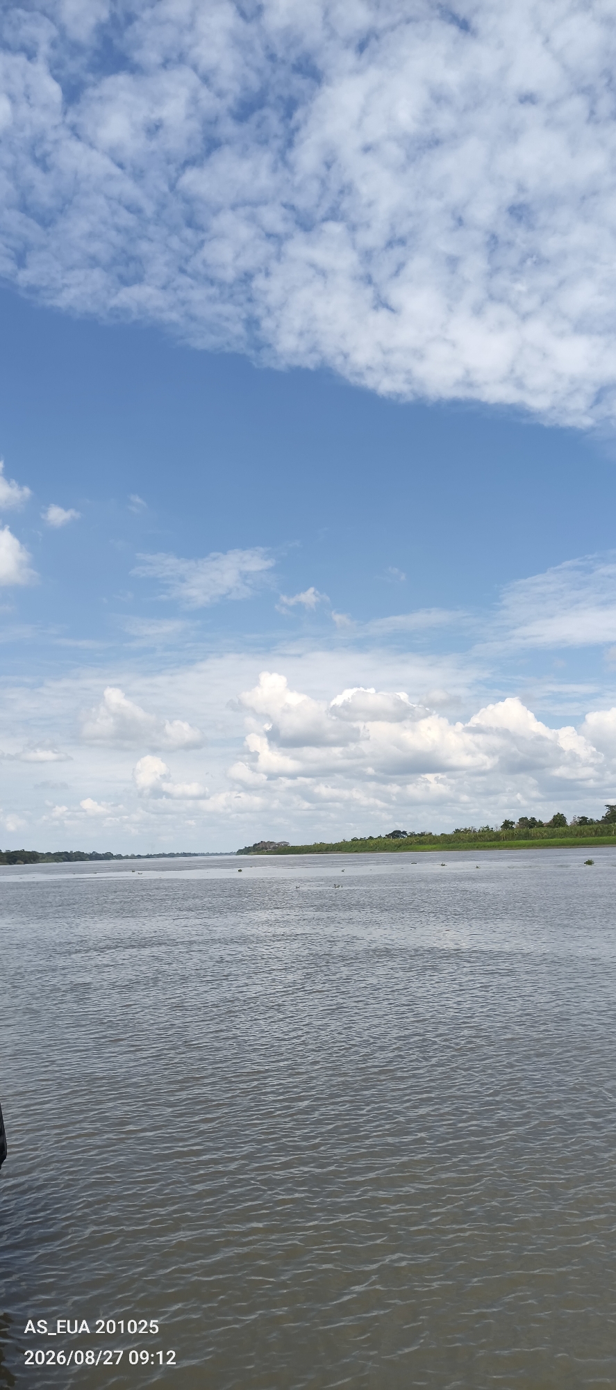 The image depicts a wide, calm river with gentle ripples on its surface. The water appears murky, reflecting the overcast sky above. The far bank of the river is lined with a dense, green treeline and some low-lying vegetation. In the distance, faint structures can be seen on the bank, suggesting human settlement. The sky is a pale blue, dominated by large, fluffy white clouds, some of which are partially obscuring the sun. The scene is tranquil and appears to be a natural landscape. The timestamp in the bottom left corner indicates "AS_EUA 201025 2026/08/27 09:12", which might relate to the location of Kanduanum 2, Papua New Guinea, though the image itself provides no direct visual cues to confirm this specific location.