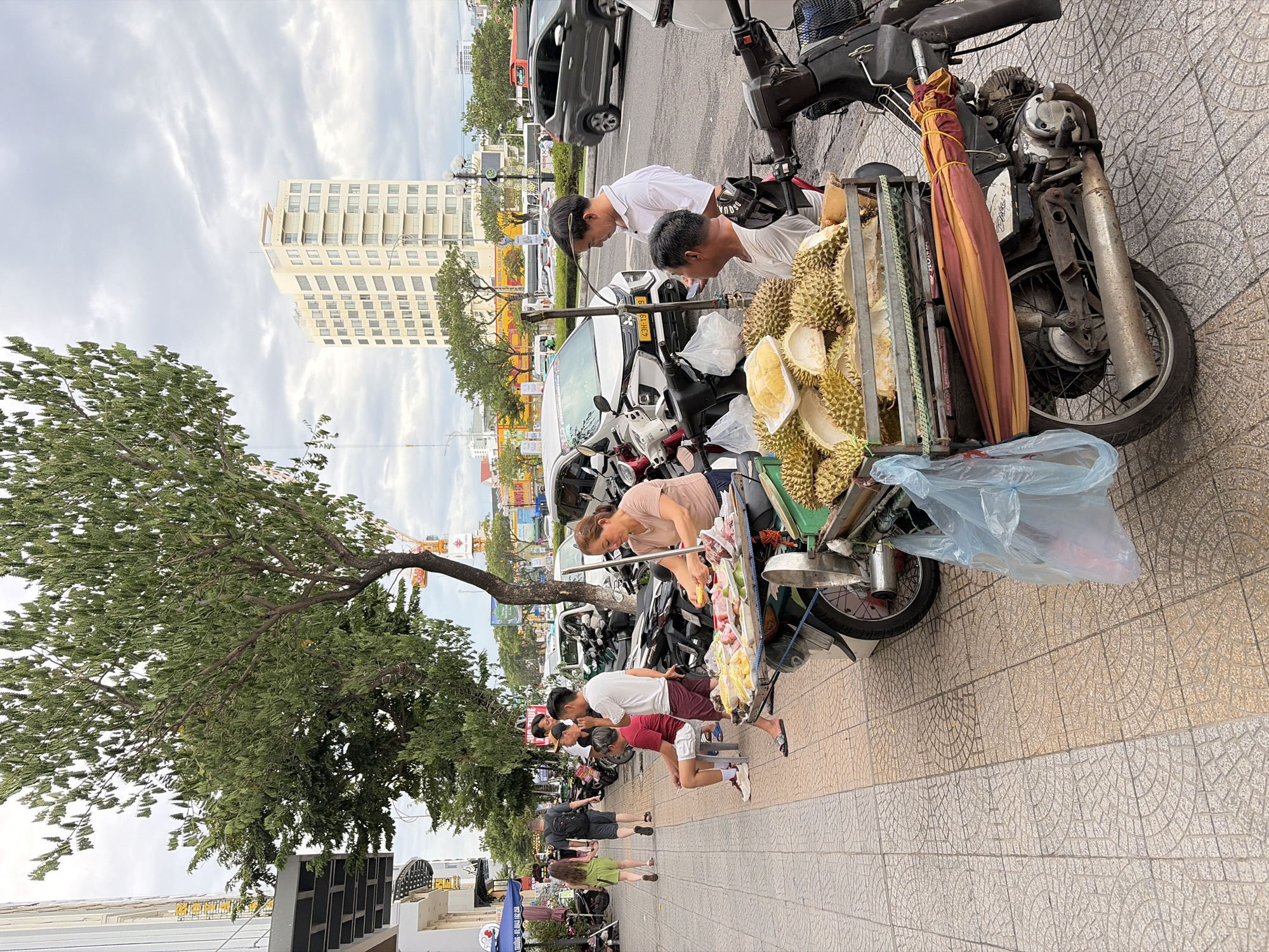 The image depicts a street scene in Da Nang, Vietnam, with a focus on a fruit stand operating from a motorbike. The fruit stand displays a large quantity of durians and other fruits, with a woman actively preparing or selling them. Several people, including customers and vendors, are present around the stand. The background shows a street with passing vehicles, a tall building, and trees, indicating an urban environment. The overall atmosphere is that of a typical street market in Vietnam.