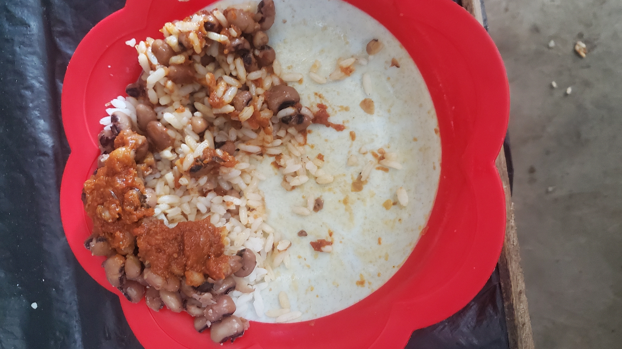 A close-up overhead shot shows a red plastic plate containing a meal of white rice and black-eyed peas. The food is partially eaten, with some of the rice and peas scraped to the side. A spicy red sauce is visible on top of the rice and peas. The plate is set on a dark surface, possibly a table, with a wooden edge visible at the bottom. The scene appears to be an outdoor or simple indoor setting, consistent with a food stall or home meal in Jalingo, Nigeria.