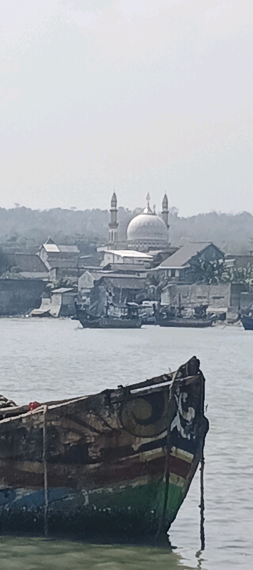 The image depicts a waterfront scene with a mosque in the background and a boat in the foreground. The mosque, a prominent structure, features a large dome and two minarets. It is situated within a cluster of buildings that form a settlement. In the middle ground, several boats are docked or anchored in the water, suggesting a harbor or fishing village. The foreground is dominated by the bow of a weathered, wooden boat. The boat is painted with various colors and appears to have decorative elements. The water is calm, reflecting the hazy sky. The overall atmosphere is one of a coastal town or village, likely in Indonesia, given the architectural style of the mosque. There are no visible people in the frame. The location is identified as Blimbing, Indonesia, suggesting this view is characteristic of the waterfront areas in that region.