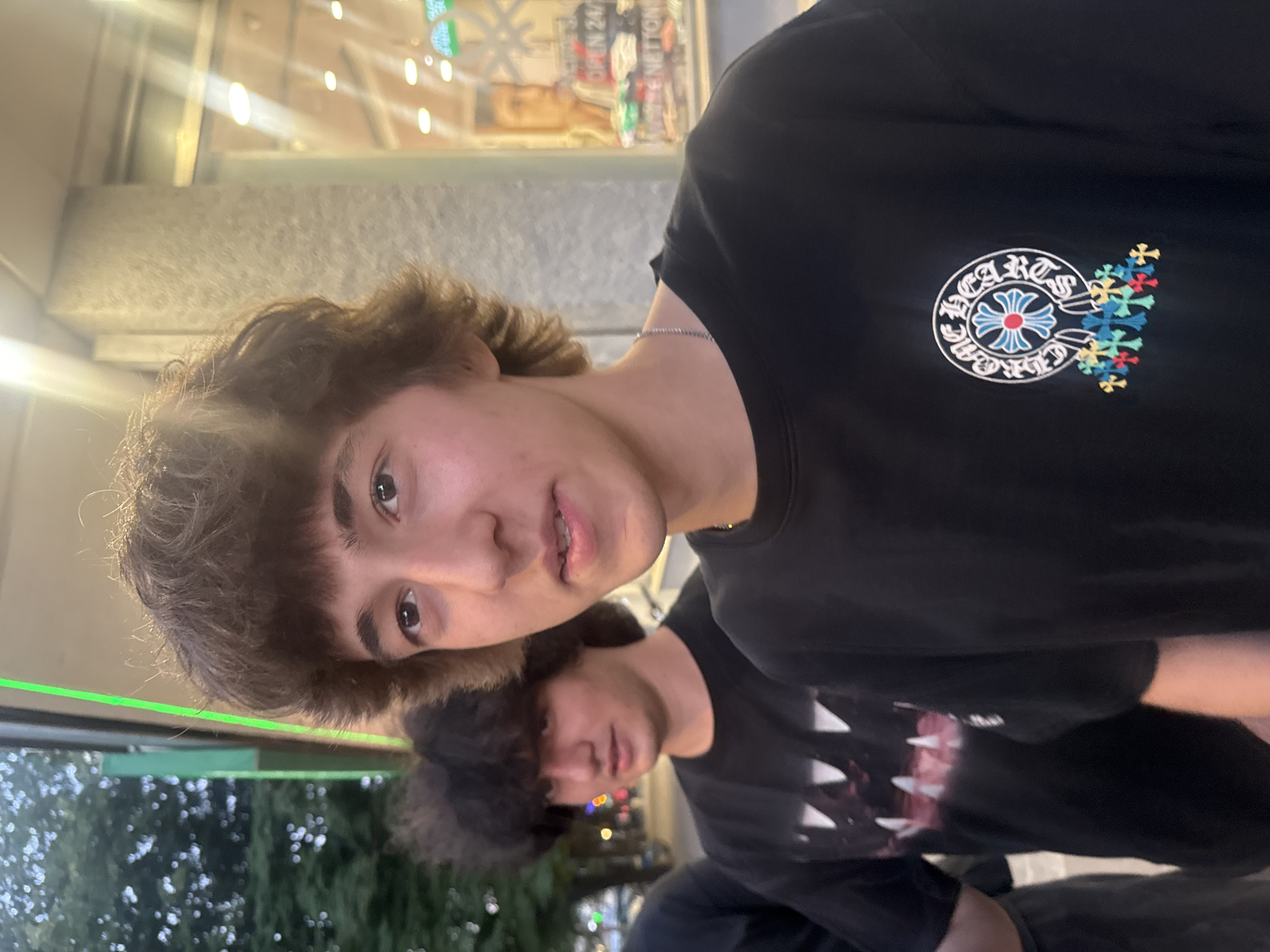 Two males are visible in an outdoor setting. The foreground male has brown hair, light skin, and is wearing a black t-shirt with a Chrome Hearts logo on the right chest. The background male also has dark hair and is wearing a black t-shirt. The background appears to include trees and urban elements with a building showing "OPEN 24" sign. The location is suspected to be Podgorica, Montenegro.