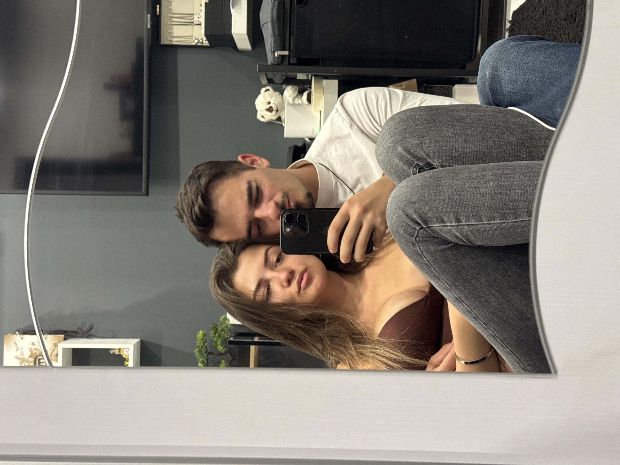 The image shows a selfie taken in a mirror. A man with short brown hair and a white shirt is holding a phone, presumably taking the picture. Beside him is a woman with long brown hair and a brown top. Both are partially visible in the reflection. The background features a gray wall, a TV, shelving with items including a teddy bear, and a decorative miniature tree. The location is in Sofia, Bulgaria.