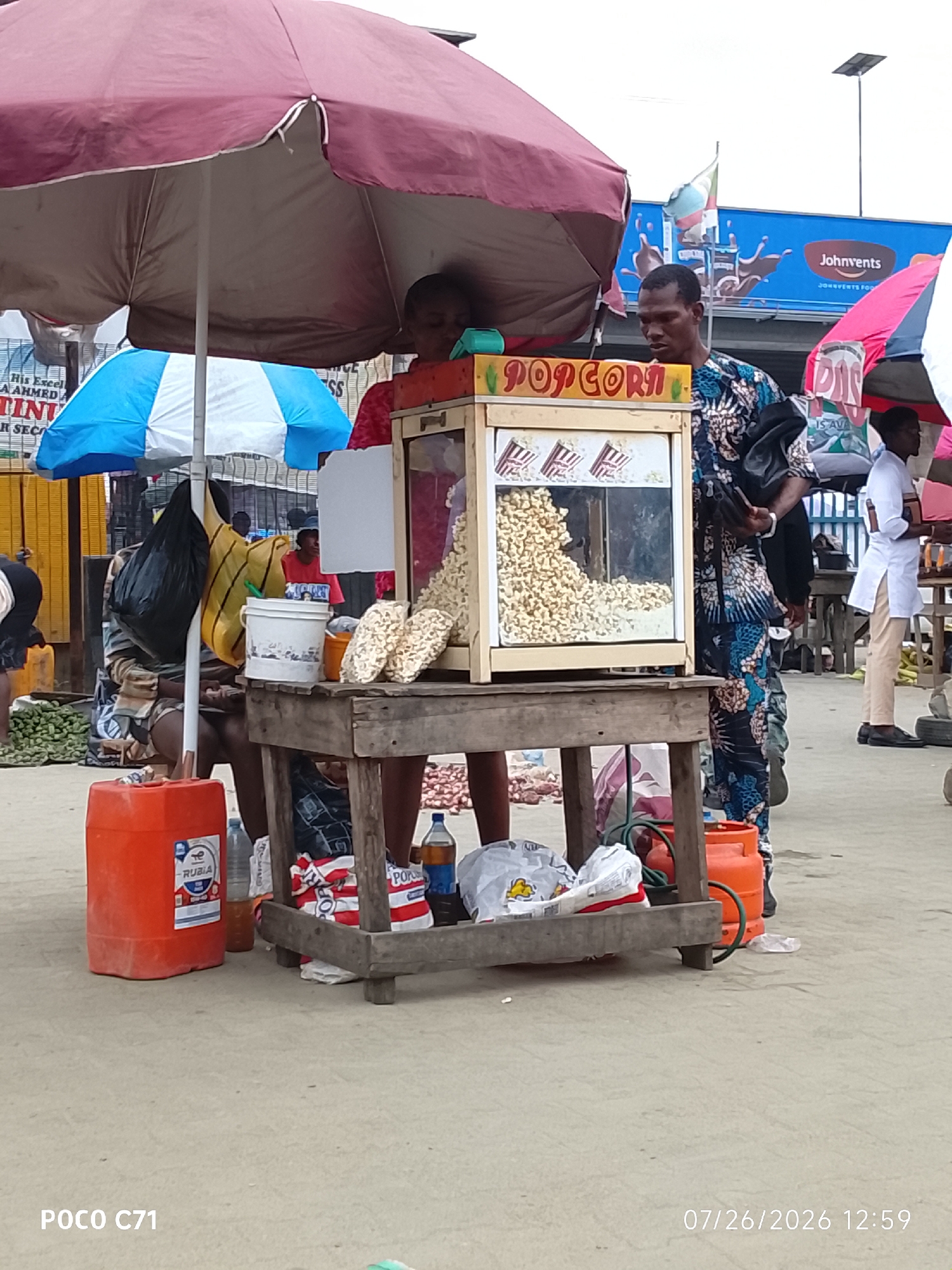 A street vendor in Lekki, Nigeria, is selling popcorn from a mobile cart under a large maroon umbrella. The cart has a glass display filled with popcorn, and the words "POPCORN" are visible in red lettering. A man wearing a colorful patterned shirt stands behind the cart, and another person in a white uniform stands to the right. Several other individuals are partially visible in the background. Various items, including large orange containers, bags of popcorn, a white bucket, and a gas cylinder, are scattered around the cart. In the background, a billboard with the "JohnVents" logo and an image of splashing chocolate is visible, suggesting the location is a commercial area. The ground appears to be paved with a light-colored surface, and the sky is overcast. The image also displays the text "POCO C71" and "07/26/2026 12:59" at the bottom.