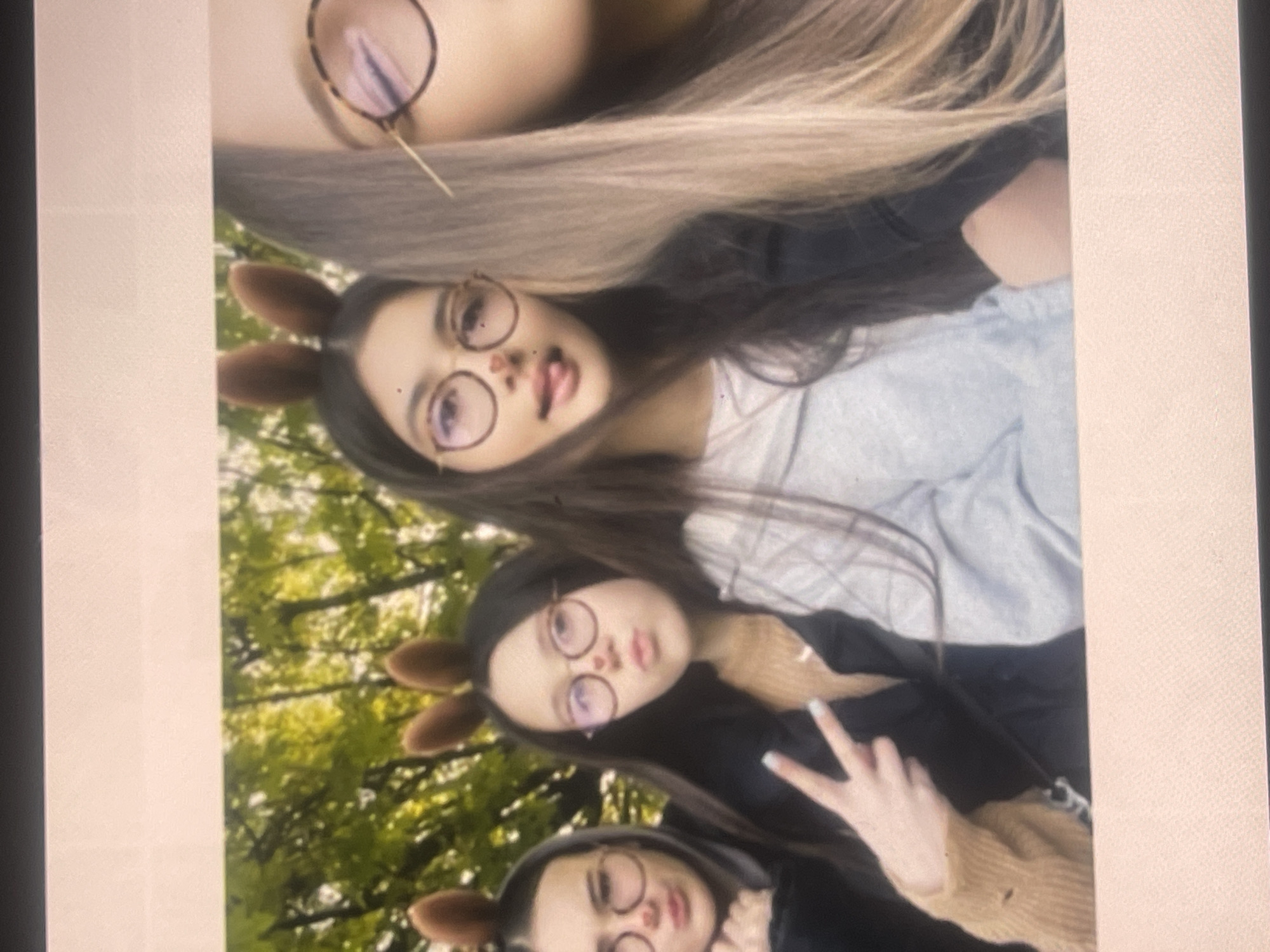 A group of four young women or girls is captured in a casual outdoor photo, likely in a park or wooded area in Poltava, Ukraine, during daylight hours. The image is oriented vertically and appears to be a selfie or a group shot.
All four individuals have long hair; three have dark hair, and one, partially visible at the top, has lighter brown or blonde hair. They are all wearing round-framed eyeglasses. Three of them are adorned with brown animal-ear headbands, resembling deer or bunny ears.
The second person from the top, with dark hair and animal ears, is playfully sticking her tongue out and is wearing a light-colored top. The third person, also with dark hair and animal ears, is making a peace sign with her left hand, which has light-colored nail polish. She is wearing a dark top underneath a light brown knitted garment. The uppermost and lowermost individuals are only partially visible, showing their faces and hair.
The background features blurred green foliage and trees, indicating a natural outdoor setting. The overall atmosphere of the photo is friendly and lighthearted, with the subjects appearing to be in good spirits. There is no visible text within the image. The picture's quality is slightly soft, possibly suggesting it is a photograph of a screen.