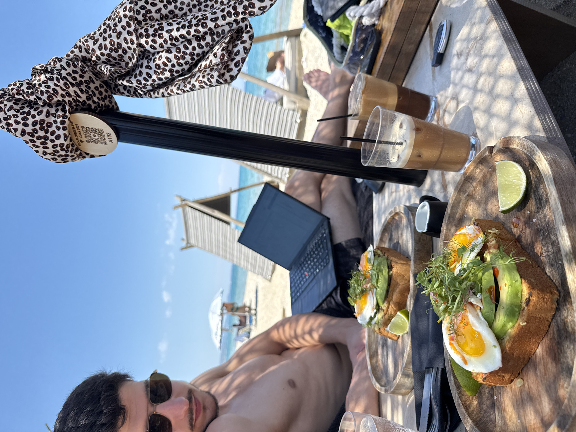 A man is relaxing on a beach in Pallini Municipal Unit, Greece, with a laptop on his lap. He is wearing sunglasses and appears to be enjoying a meal of avocado toast with fried eggs and a drink. In the background, beach chairs and umbrellas are visible, along with the ocean and a clear blue sky. A patterned scarf is tied to a black pole in the foreground, with a QR code attached to it. The table also holds two glasses of iced coffee and a small white cup.