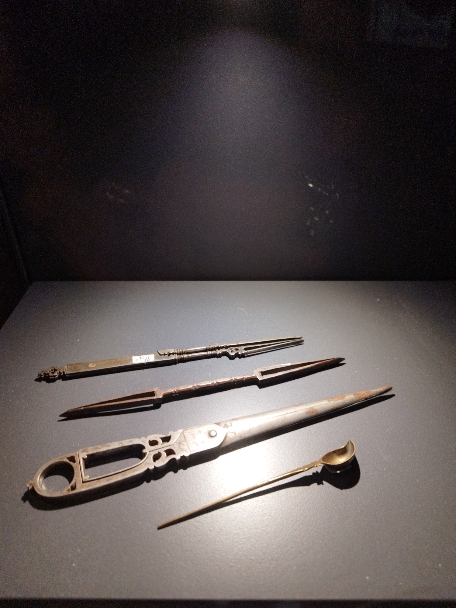 An exhibit case contains four antique, metallic utensils arranged horizontally on a dark gray, matte surface. The uppermost utensil is a long, slender object with decorative finials at each end and a central segmented body. Below it, a second utensil is visible, featuring a pointed, bifurcated end and a rectangular channel in its midsection. The third item is a complex implement with a large, circular loop at the left and a more ornate, carved handle to the right. This object has an opening in its center and decorative cutouts. Finally, the lowest and smallest object is a delicate spoon with a shallow, oval bowl and a long, thin handle, resting at an angle. The background is a dark, shadowed wall.