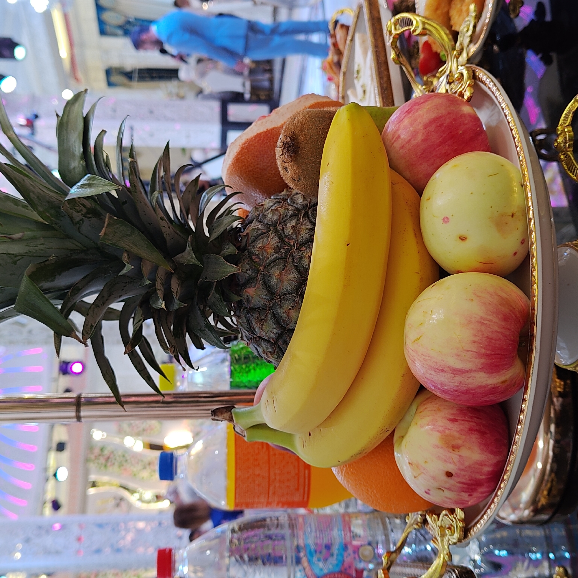A fruit arrangement is displayed on a tiered serving stand, likely at a gathering or event in Khujand, Tajikistan. The arrangement features a pineapple at the top, with a bunch of bananas below it. To the side of the bananas, there is a halved grapefruit and a kiwi. In the foreground, a white dish with golden trim holds several apples, some with red and yellow streaks, and one that is mostly yellow with red spots. Behind the fruit, several bottles are visible, including a clear bottle with a blue cap, an orange-colored drink in a bottle with a red cap, and a green liquid in a bottle. The background is blurred, showing an indoor environment with people dressed in formal attire and decorative lighting, suggesting a celebration or banquet. One person in the background is wearing a blue suit. The overall scene is brightly lit and appears to be a festive occasion.