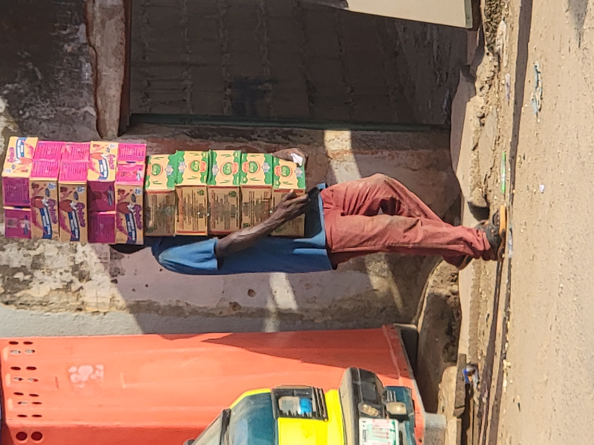 A man lies prone on a concrete surface next to a building in Maiburiji, Nigeria. His head and torso are obscured by stacks of pink and green boxed goods that appear to be his wares. He wears a blue shirt and reddish-brown pants. His legs extend along the sidewalk. The lower portion of the image contains an orange container and part of a vehicle with yellow and blue elements. The location appears to be a commercial area.