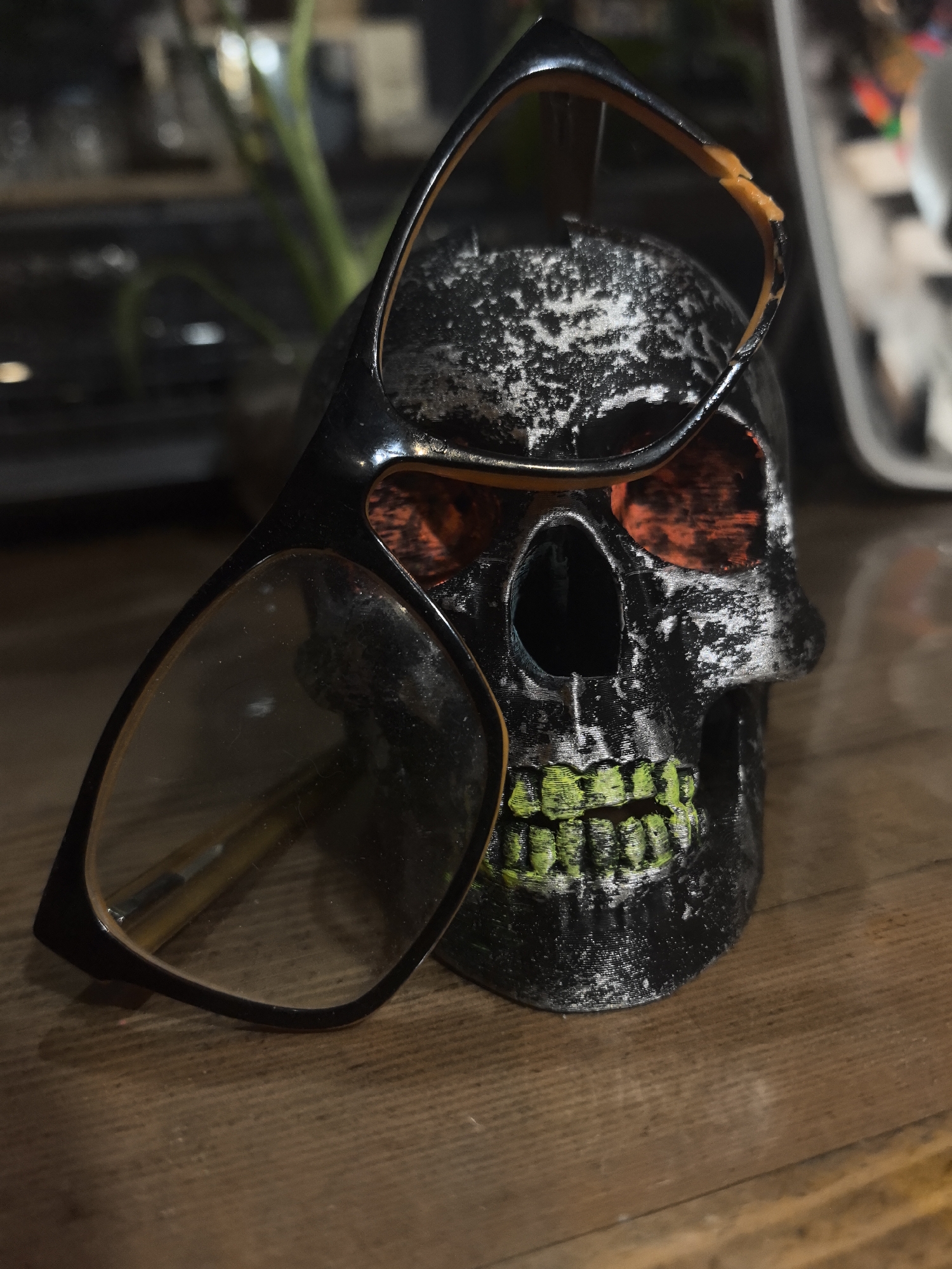 The image shows a black skull with orange eye sockets and green teeth placed on a wooden surface. A pair of glasses with a black and orange frame rests on top of the skull. The glasses appear to be damaged, with a broken section near the top of the frame. Background details include indistinct objects, possibly plants or furniture, suggesting an indoor environment. The location is not specified within the image.