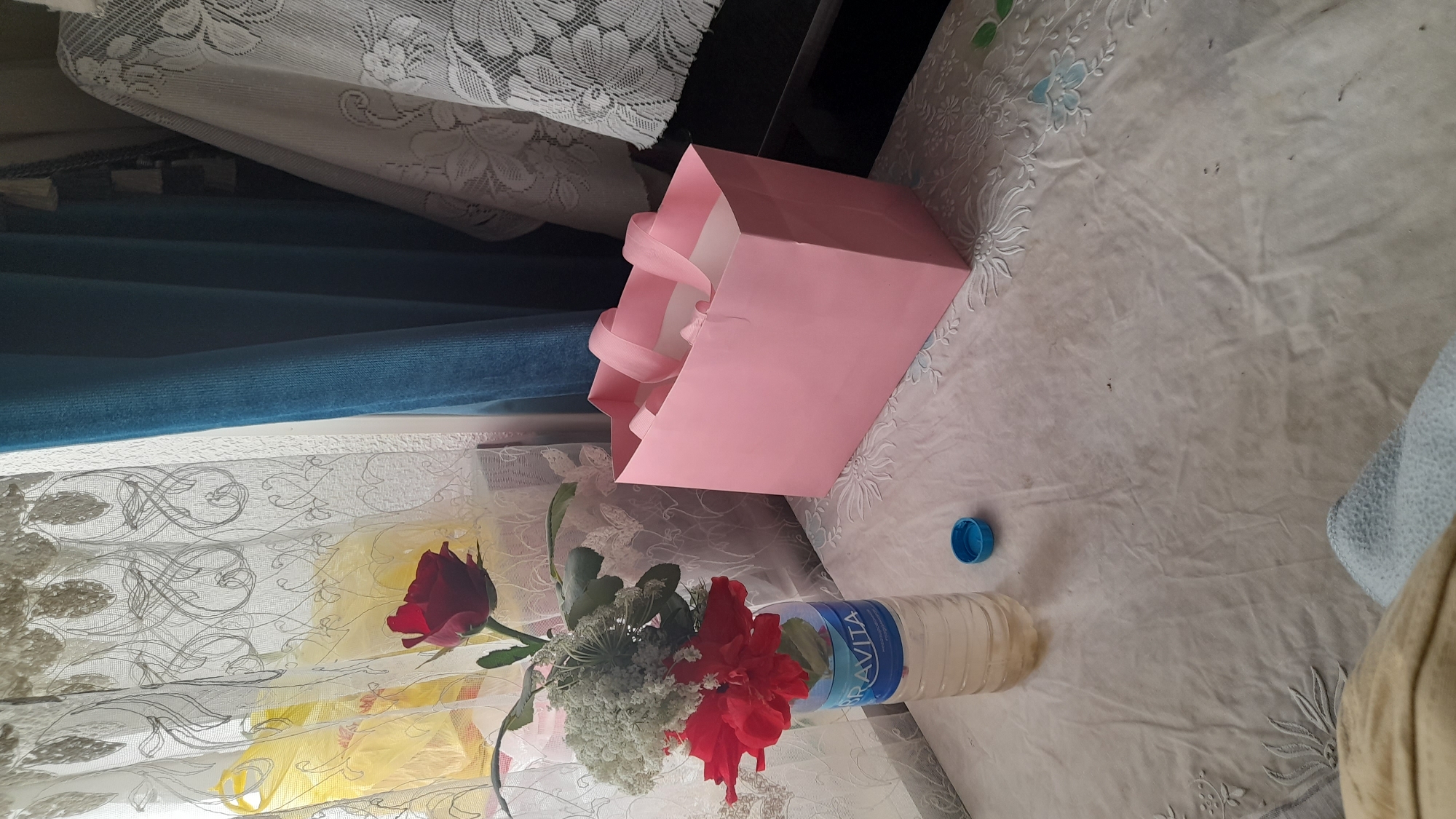 In Osh City, Kyrgyzstan, a scene unfolds on a table covered with a white, embroidered cloth. A plastic bottle, serving as a makeshift vase, holds a bouquet of red flowers, a single red rose, and a cluster of small white blossoms. Next to the bottle, a blue cap rests on the cloth. To the right, a pink gift bag with ribbon handles stands open. Behind the table, sheer white curtains with an intricate pattern frame a window, behind which a yellow plastic bag is visible. A thick blue curtain hangs to the right, partially obscuring more white fabric adorned with floral embroidery. The lighting suggests an indoor setting, possibly a home, with natural light filtering through the curtains.