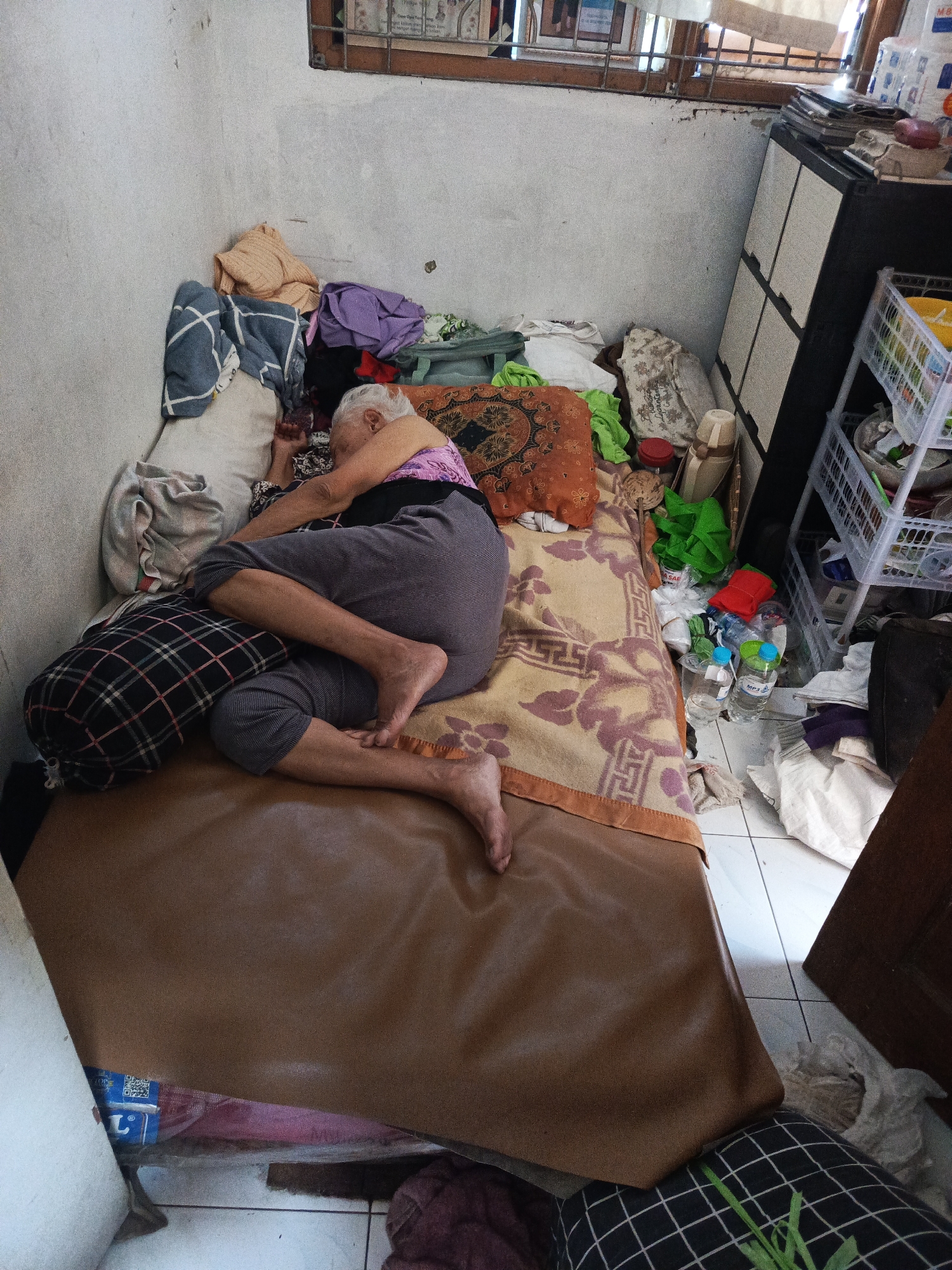 An elderly woman with white hair sleeps on a bed in a cramped room. She is lying on her side, facing away from the camera, with her legs bent. She is wearing a purple top and grey pants. The bed is covered with a brown blanket with a floral pattern and a brown leather-like sheet. There are several pillows on the bed, including one with a black and white checkered pattern. The room is cluttered with various items, including clothes, bottles of water, and a plastic shelf unit. A cabinet is visible in the background. The room appears to be a bedroom. The image was taken in Yogyakarta, Indonesia.