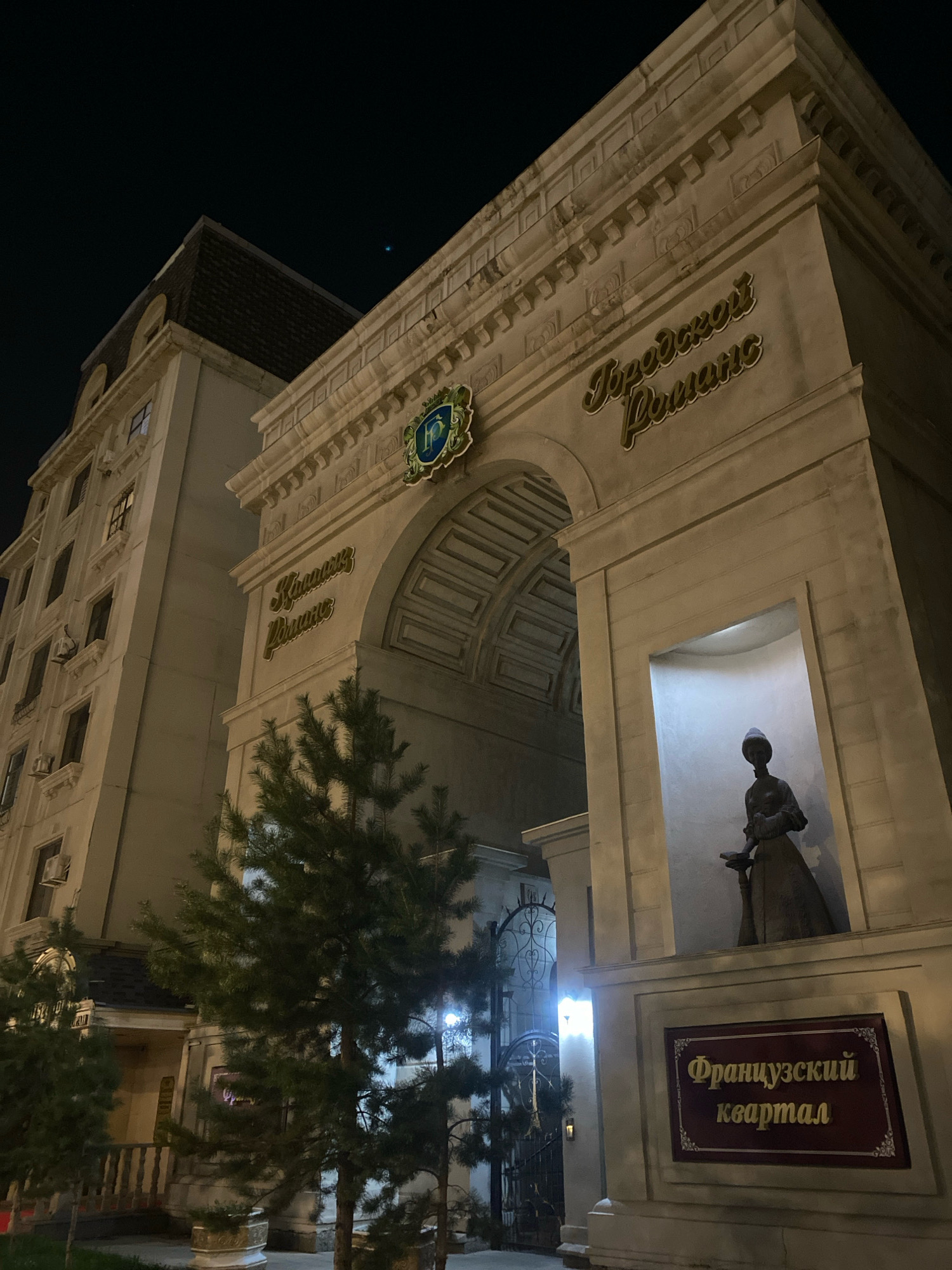 The image shows the facade of a building at night in Astana, Kazakhstan, specifically the "French Quarter" (Французский квартал) residential complex. The main subject is an architectural archway designed to resemble a triumphal arch. Above the arch, golden Cyrillic lettering reads "Городской Романс" (City Romance). To the left of the arch, a tall, multi-story building with a mansard roof is visible, illuminated by interior lights. To the right of the arch, a statue of a woman in historical dress is displayed in a recessed niche. Below the niche, a dark red sign with gold Cyrillic lettering reads "Французский квартал" (French Quarter). Pine trees are positioned in front of the structure, and an ornate metal gate is visible in the background. The scene is lit by artificial lighting, casting shadows and highlighting the architectural details.