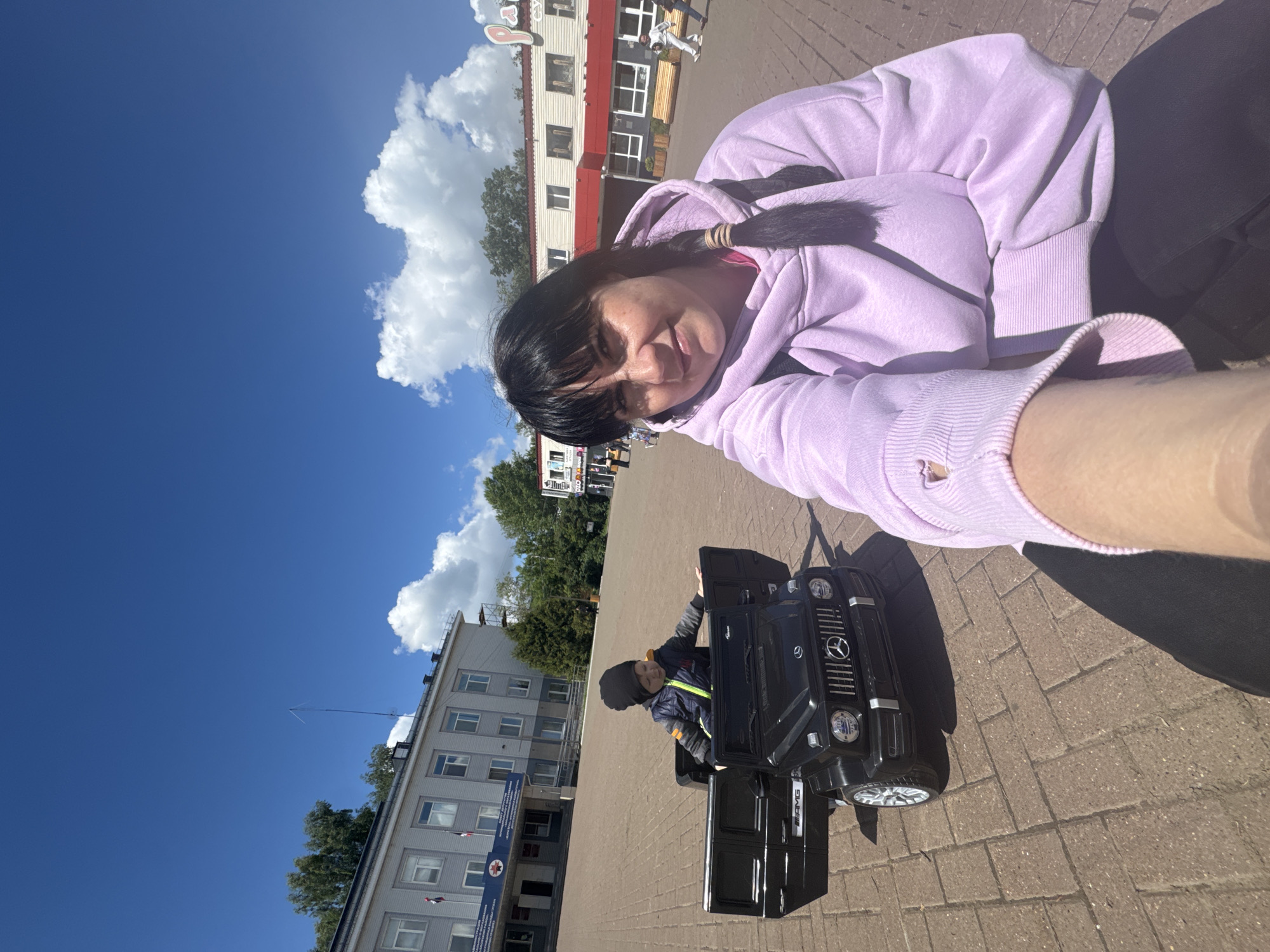 A woman and child are in a public space, presumably in Zav'yalovo, Russia. The woman is taking a selfie. She wears a light purple hoodie, has dark hair, and is positioned in the upper part of the frame. The child is seated in a black toy car, which resembles a Mercedes-Benz G-Class SUV. The car's doors are open.
In the background are buildings, including one with a red roof, and others that seem to be administrative or public buildings. The scene is brightly lit with a blue sky and some clouds overhead. The ground is paved with brick or stone.