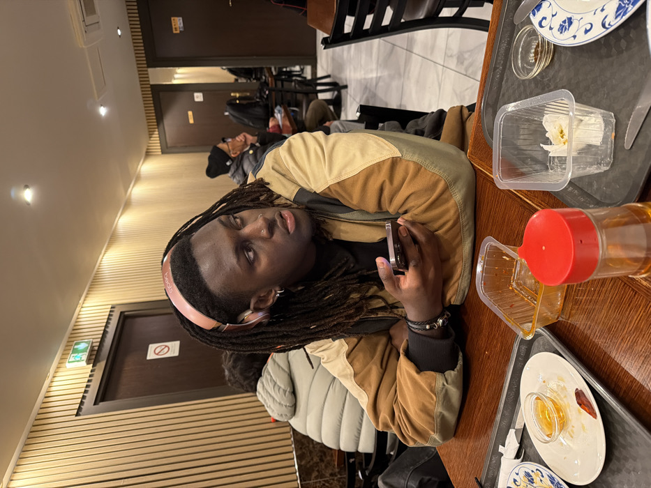 A person with dreadlocks and pink Beats headphones is sitting at a wooden table. They are wearing a brown and beige jacket and holding a smartphone. The table has trays with empty dishes, a plastic container with napkins, and a bottle with a red lid. Behind the person are other people and chairs. The background includes a wall with wood paneling and doors with signs.