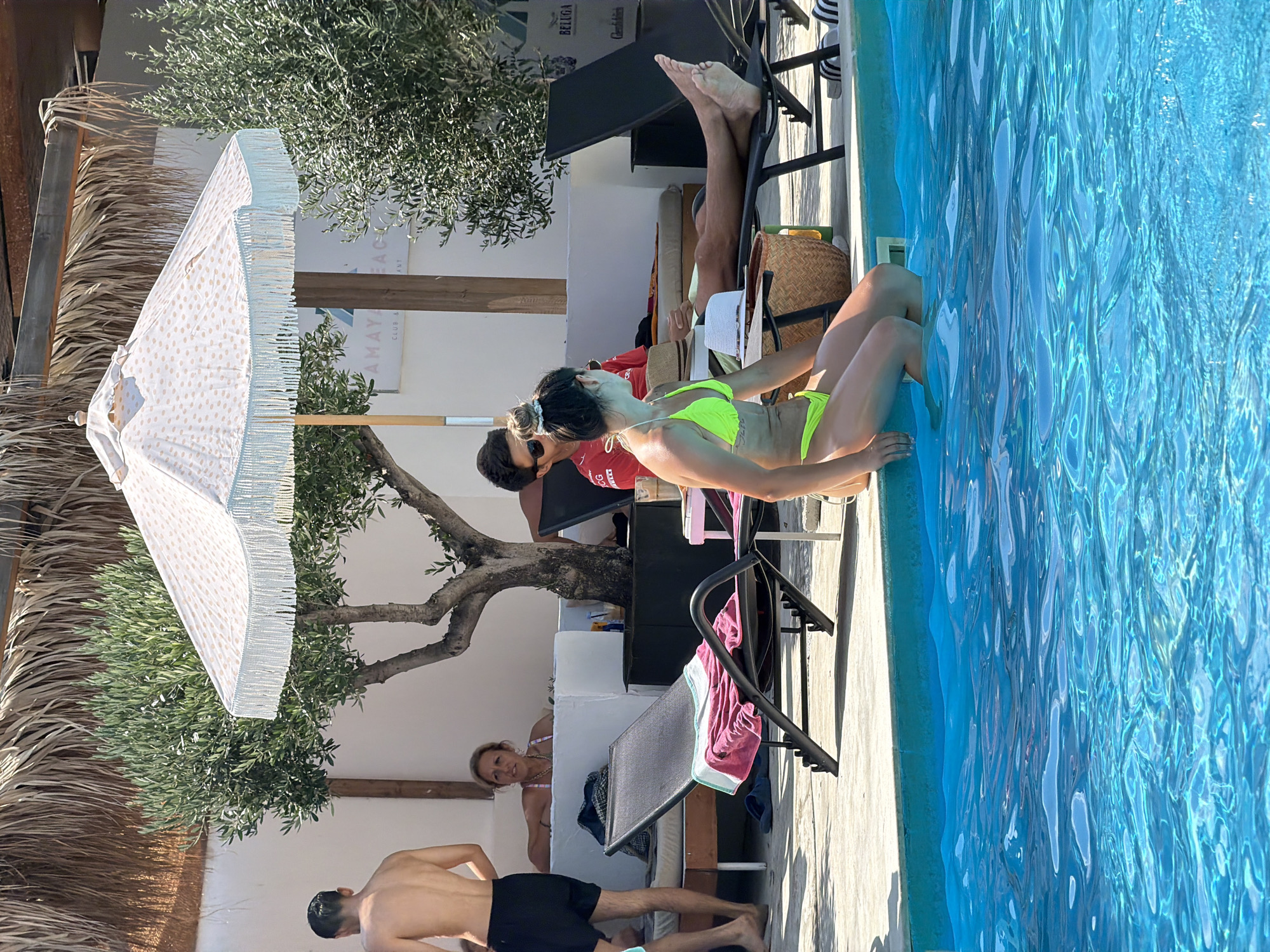 The image depicts a sunny outdoor scene at a beach club with a pool. In the foreground, a woman in a neon yellow bikini sits on the edge of the pool, her legs submerged in the bright blue water. Behind her, several people are relaxing on lounge chairs under a patterned umbrella and trees. One man, wearing a red shirt, is seated on a lounge chair. Another man, shirtless and wearing black shorts, stands near the pool. A woman with blonde hair is visible further back, seated. In the background, there is a white building with a sign that reads "AMAYA BEACH CLUB & RESTAURANT". The overall environment suggests a leisurely day at a resort or beachside establishment. The location is implied to be in Burgas, Bulgaria, based on the provided information.