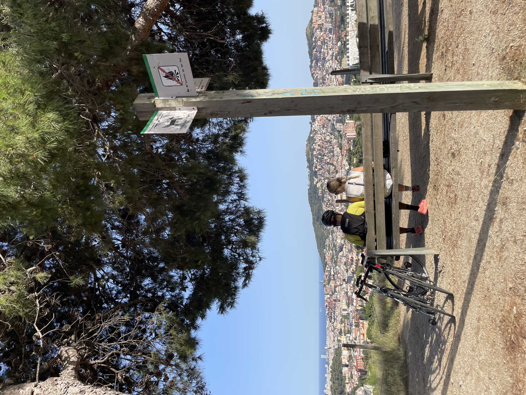 The image depicts a scenic overlook in Barcelona, Spain, offering a panoramic view of the city and the Mediterranean Sea. In the foreground, a wooden bench is occupied by two individuals, a man and a woman, both facing away from the camera and towards the cityscape. The man is wearing a bright yellow shirt and a black backpack, while the woman is dressed in a white top. Two bicycles are parked beside the bench, leaning against each other. A wooden post stands to the right of the bench, adorned with two signs. One sign, with a red circle and a diagonal line through a flame, indicates that fires are not permitted. The other sign, featuring a silhouette of a dog, likely relates to pet regulations. The background reveals the sprawling urban landscape of Barcelona, characterized by a dense collection of buildings and hills, with the blue expanse of the sea visible in the distance. Lush green trees provide shade in the upper portion of the frame, suggesting a park or natural reserve setting within or on the outskirts of the city. The sky is clear and bright blue, indicating a sunny day.