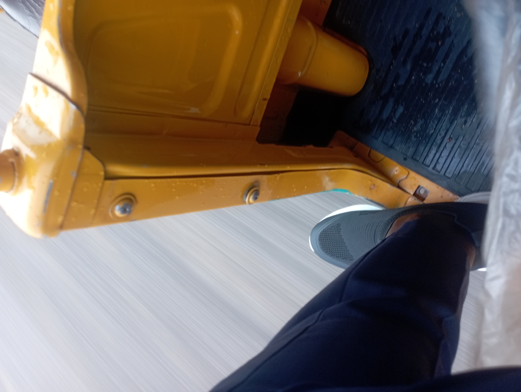 The image shows a close-up, low-angle shot from inside a yellow auto-rickshaw, likely a commercial passenger vehicle. The viewer's left leg, clad in dark blue trousers and a grey sneaker, is prominently featured, suggesting the perspective of a passenger or driver. The auto-rickshaw's yellow painted metal body, showing signs of weathering and water droplets, occupies the right side of the frame. The interior floor is a dark, ridged rubber mat. The background is a blur of grey and white streaks, indicating motion, typical of a vehicle traveling at speed on a paved road in Jos, Nigeria. The scene suggests a common mode of transportation within the city, possibly during or after rainfall, given the water droplets on the vehicle.